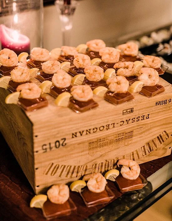 Assorted shrimp and chocolate dessert squares with lemon wedges on top, arranged on a wooden box at a celebration event.