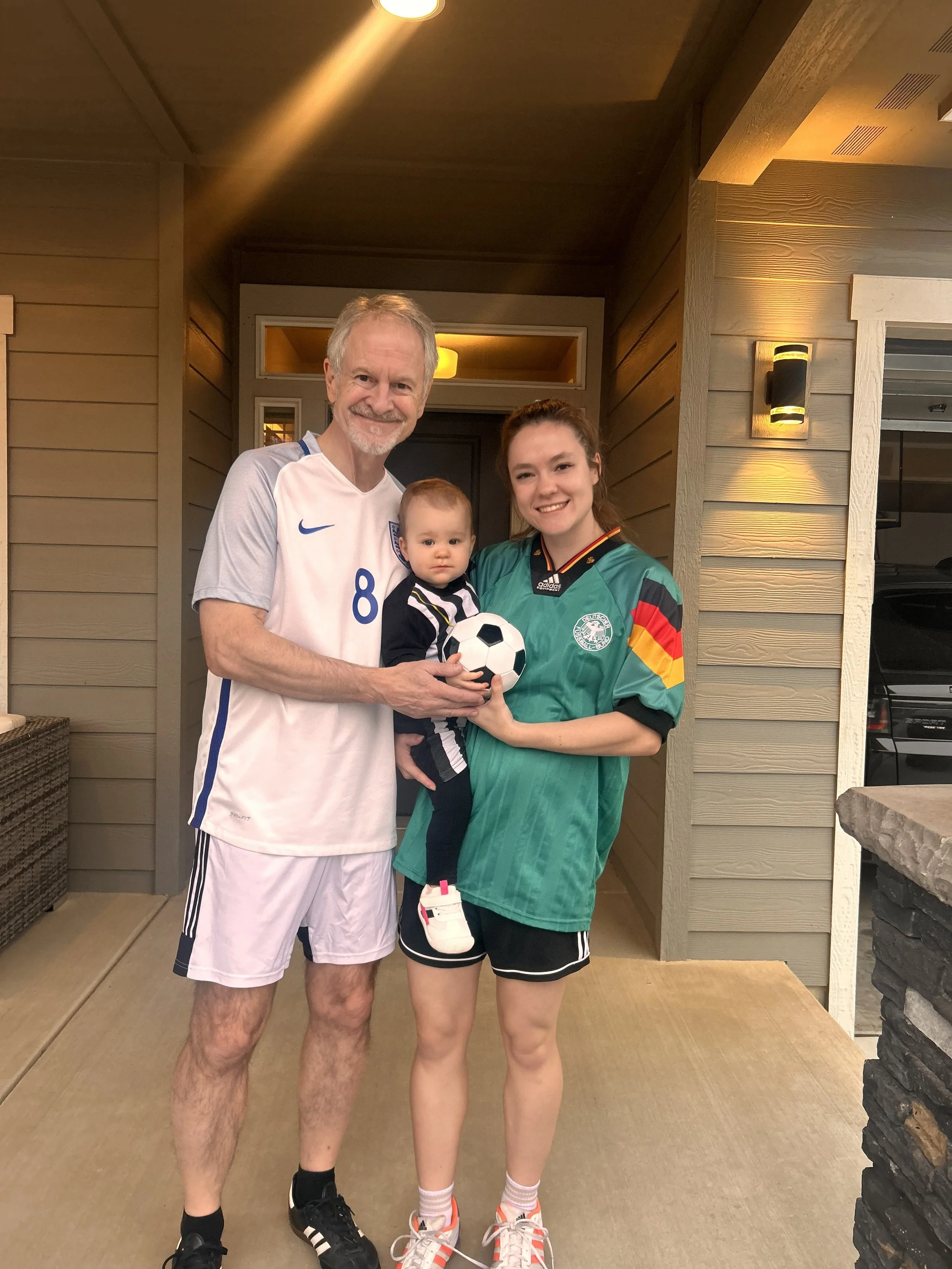 Abby Sprague and her family standing together outside a house, holding a soccer ball. The man is dressed in a white athletic jersey and shorts, the woman in a green sports jersey and black shorts, and the baby in a ref's costume.