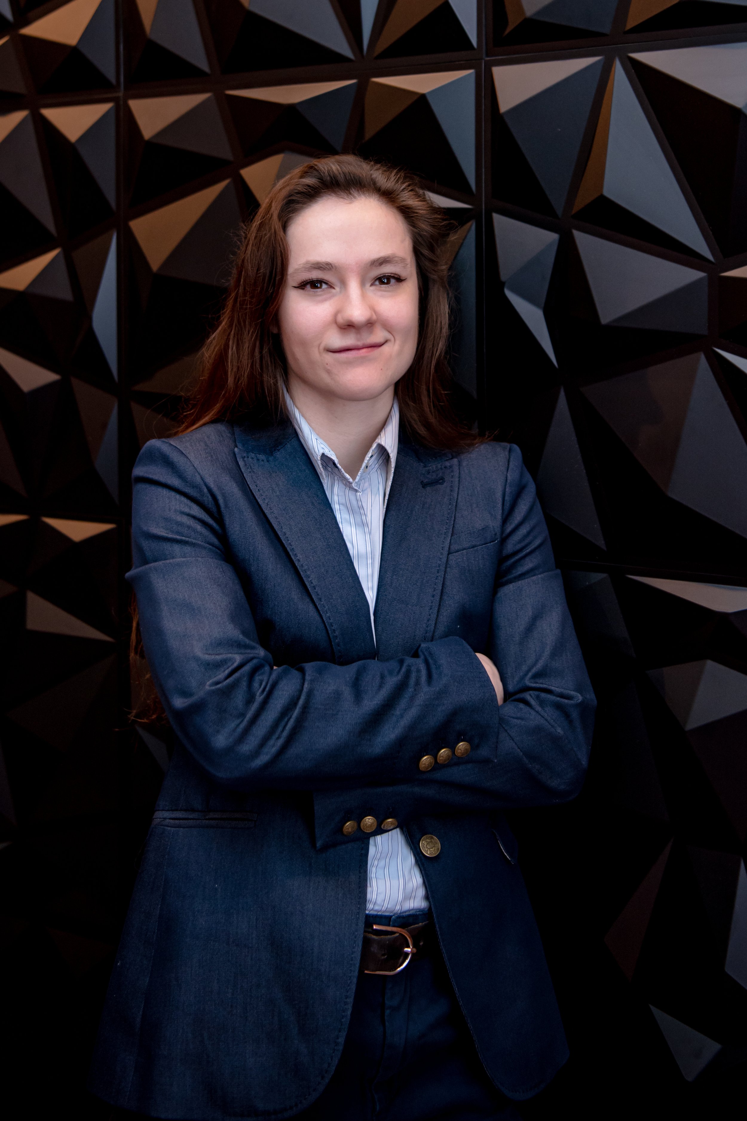 A woman with long brown hair wearing a navy blazer and light blue shirt standing with arms crossed in front of a black geometric wall.