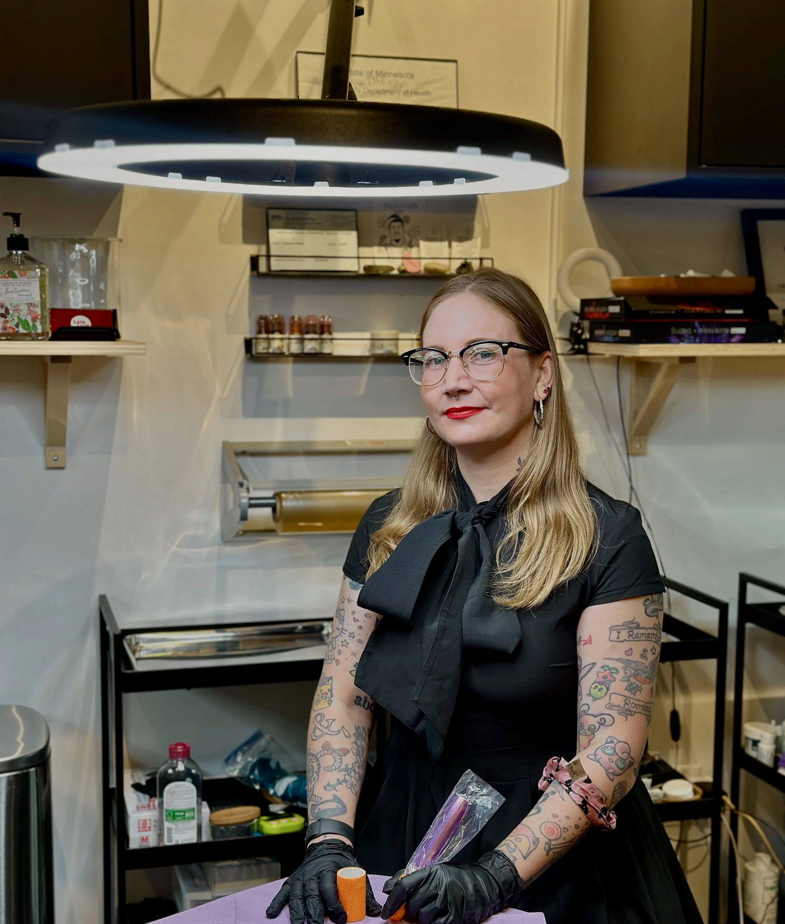 A woman with tattoos and glasses standing in a room, wearing a black dress with a big bow tie, holding a wrapped object and an orange roller, under a large round light fixture.