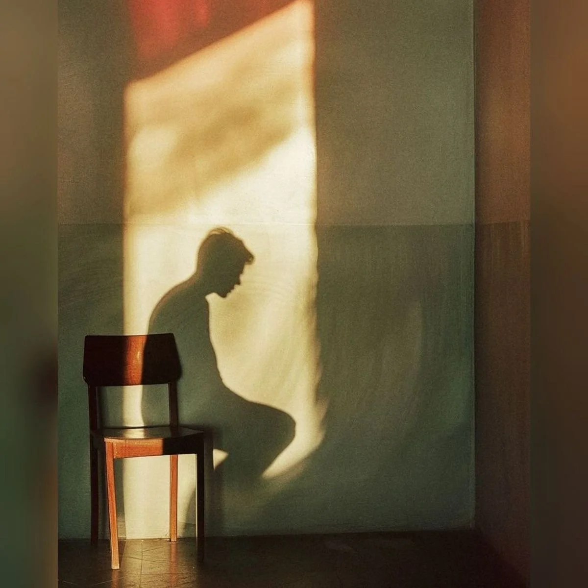 A shadow of a male-presenting person who looks like he is sitting in a brown chair that is leaned against the wall his shadow is cast on.