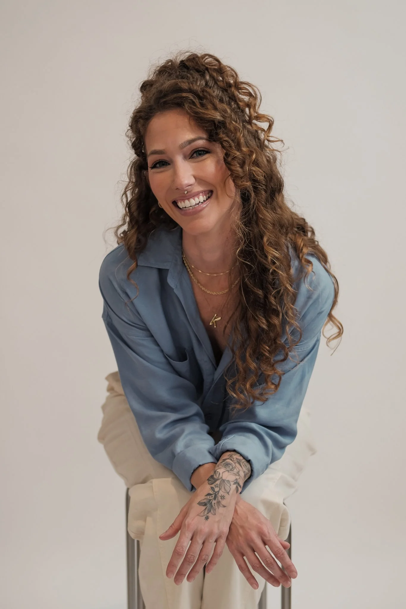 Picture of a female-presenting white person with curly hair, wearing a blue button up and khaki pants, smiling and looking at the camera