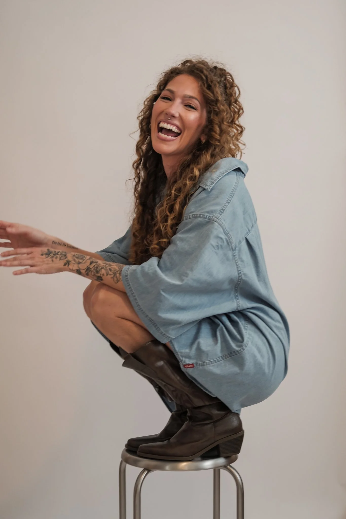 A white, curly-head, female-presenting person squatting on a silver stool in cowboy boots and an oversized button up, laughing.