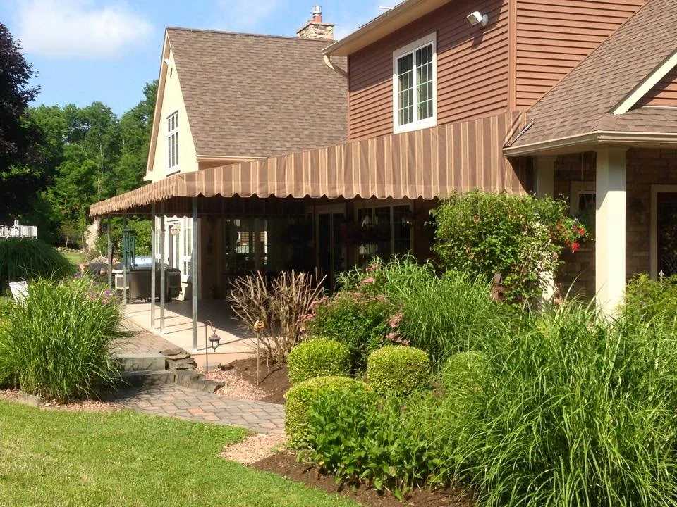 A house with a brown wooden exterior and a sloped roof, featuring a screened-in porch with a striped awning, surrounded by a landscaped garden with green shrubs, flowering plants, and grass.