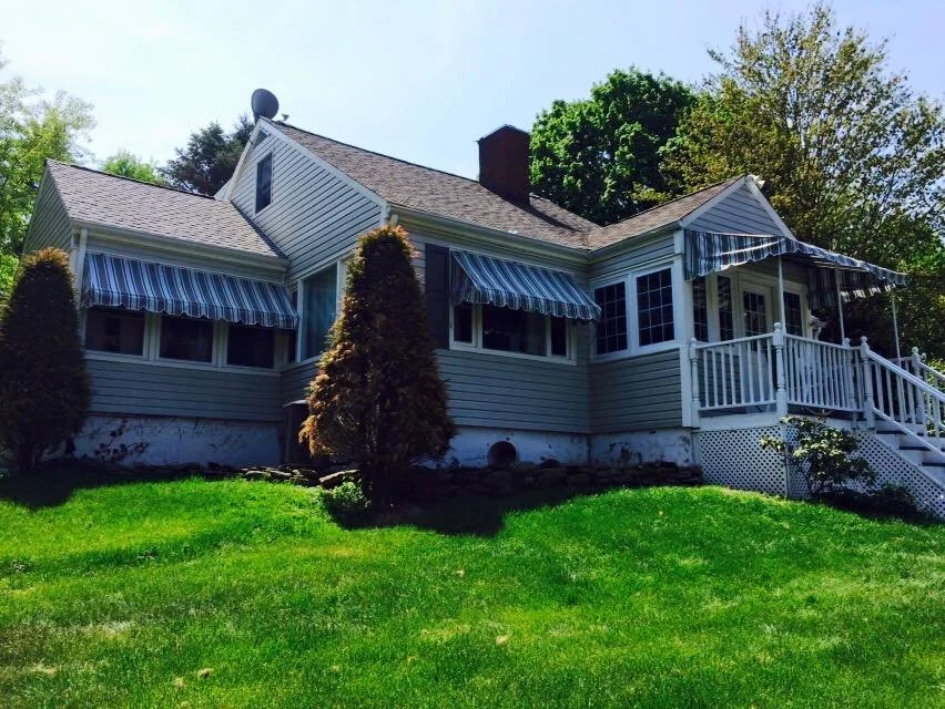 White house with a porch, striped awnings, and surrounding greenery.