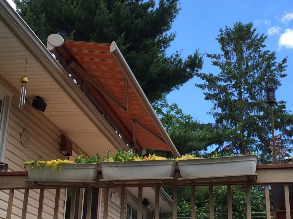 View of a house's balcony with three large rectangular flower boxes filled with colorful flowers, a brown adjustable patio awning, and tall trees in the background under a blue sky.
