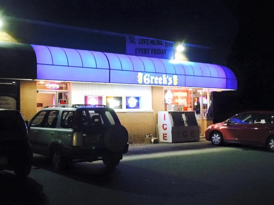 Night view of a small restaurant with a purple awning and bright neon signs, parked cars in front, and a sign indicating live music every Friday.