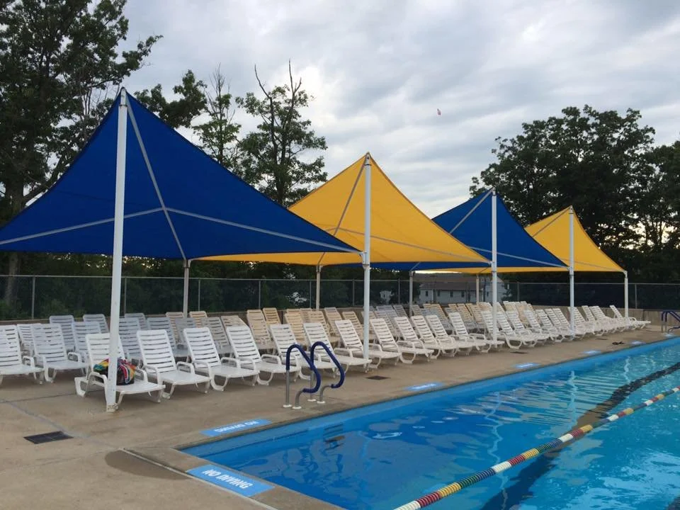 Swimming pool with white lounge chairs and yellow and blue shade tents along the poolside on a cloudy day.