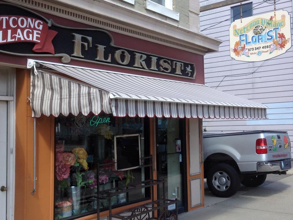 A flower shop named 'Floris' with a red and black sign, striped awning, and a window display of flowers. Next to it is a floral delivery sign and a parked white pickup truck.