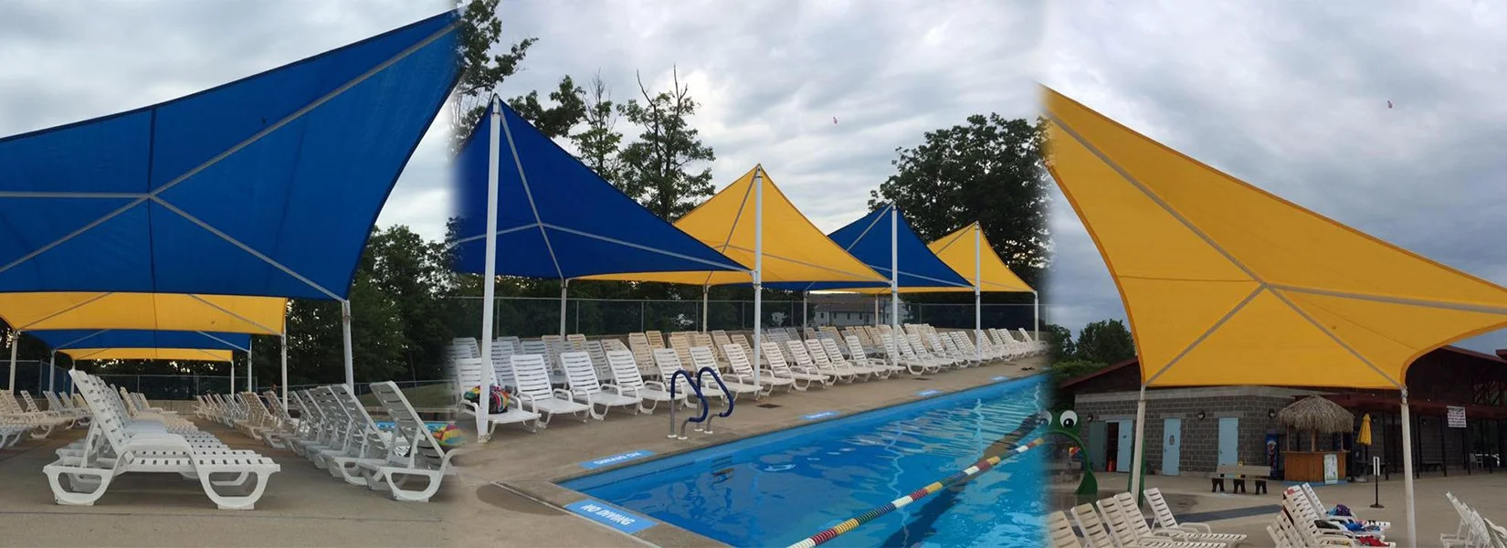 Poolside area with empty white lounge chairs and large blue and yellow shade sails over the pool and seating area, with trees and cloudy sky in the background.