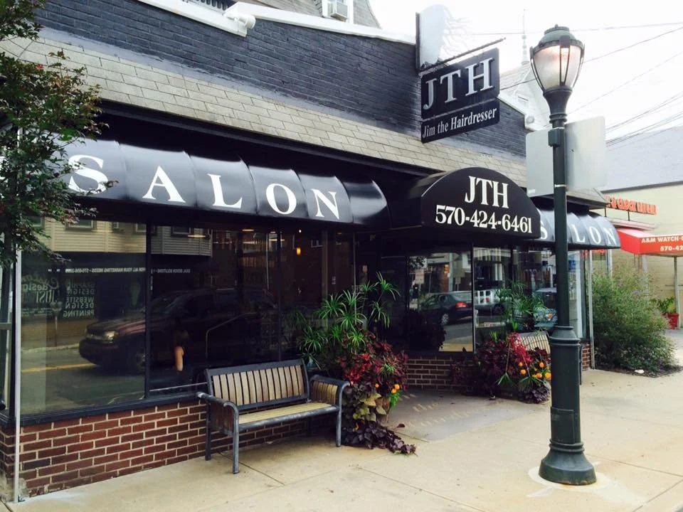 Exterior of a hair salon with a black awning displaying 'SALON' in white letters, a sign 'JTH Jim the Hairdresser' with contact number, a streetlamp, and seating area with plants and flowers outside.