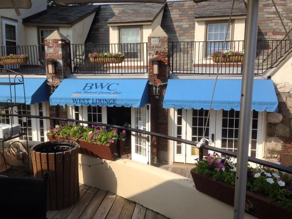 Outdoor balcony with blue awnings, flower boxes with pink and white flowers, brick and stone building, and a sign that reads 'BWC West Lounge'.