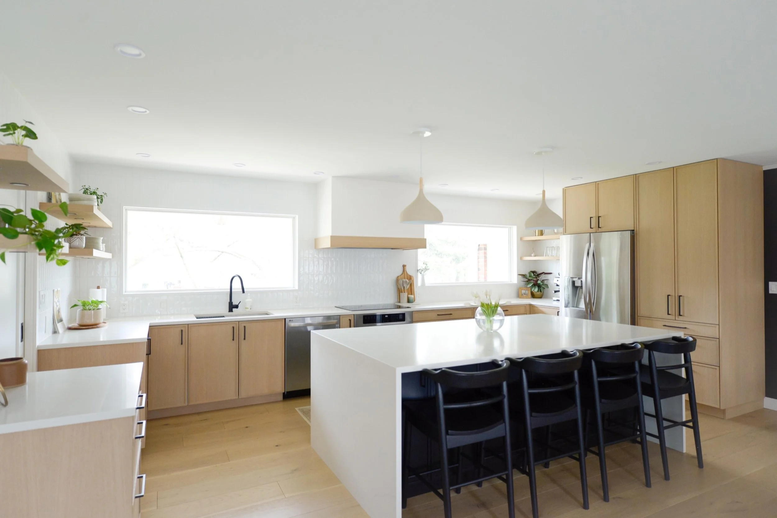 Bright modern kitchen with light wood cabinets, a large white island with four black chairs, stainless steel refrigerator, and two pendant lights hanging from the ceiling.