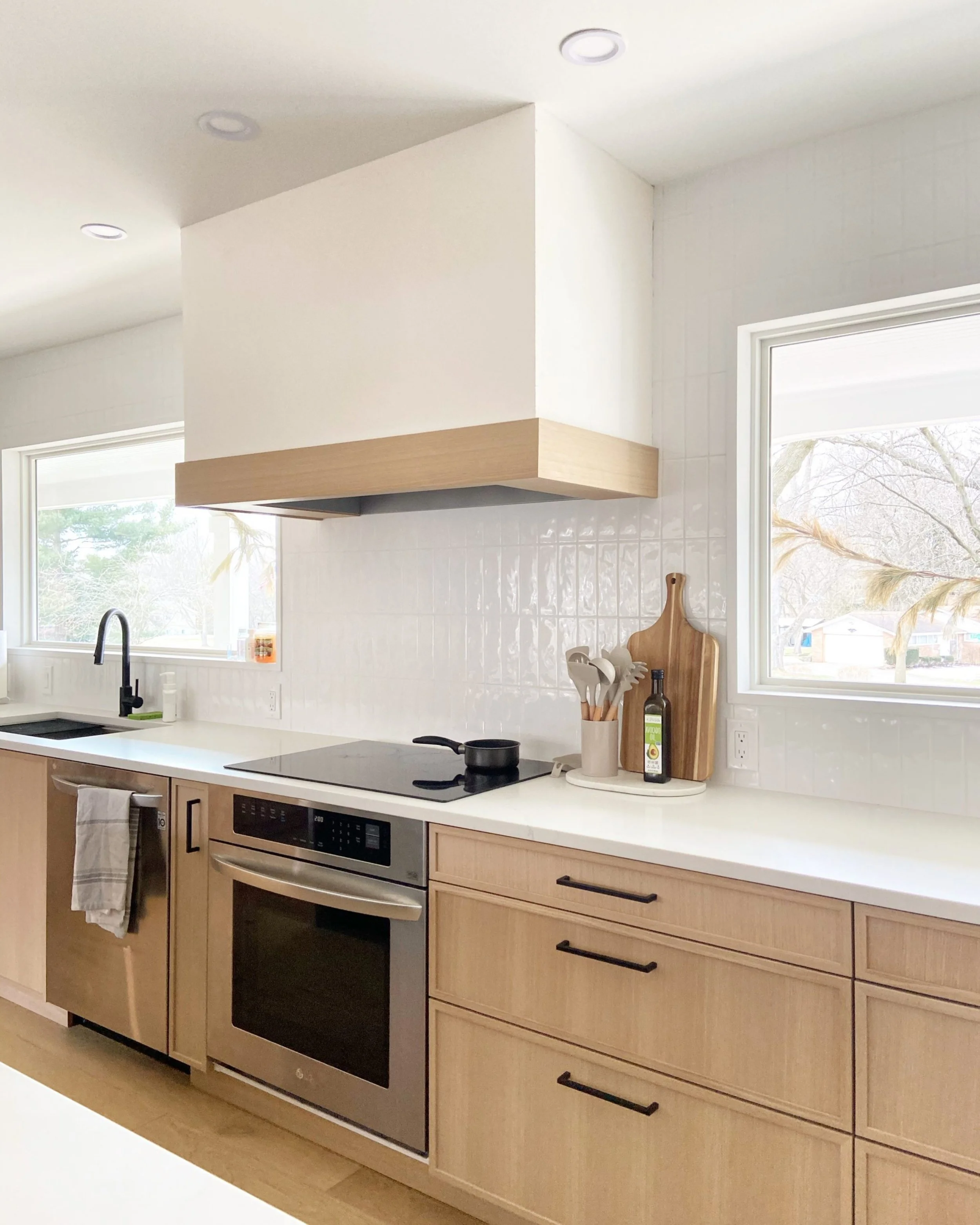 Modern kitchen with light wood cabinets, white countertop, black sink, and windows with natural light.