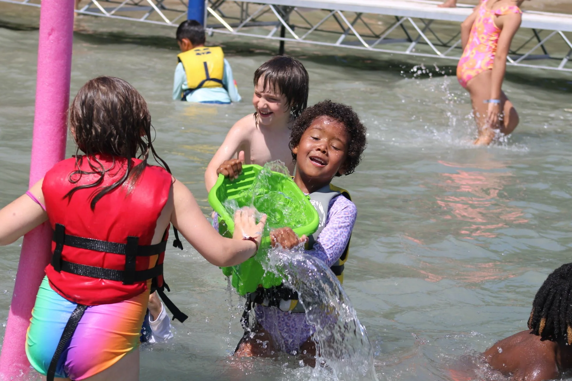 Children enjoying a water fight in a pool, with some kids splashing water, wearing life jackets and colorful swimsuits.