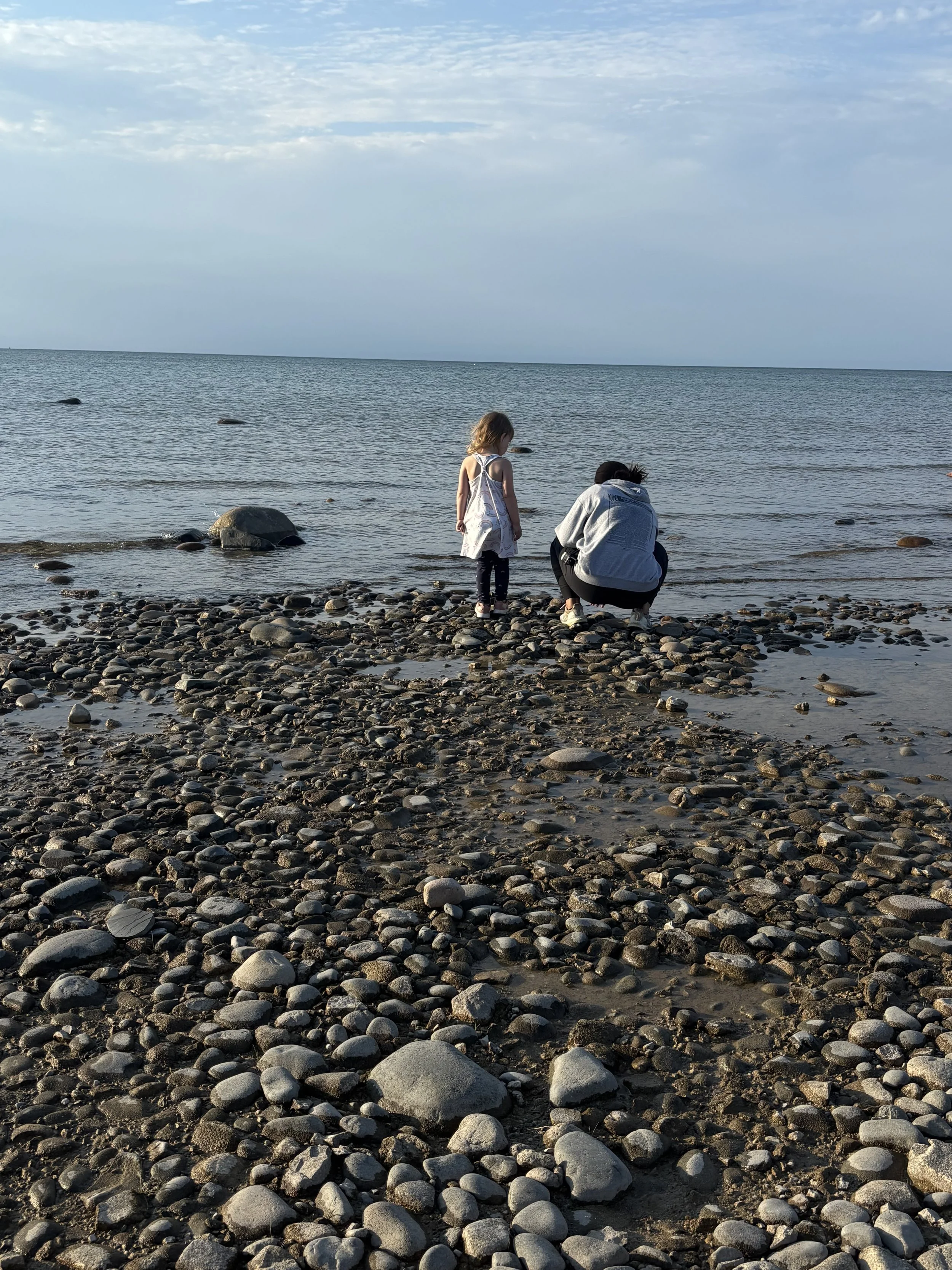 A woman and a young girl on a pebbled beach near the water, with the woman squatting and the girl standing, both facing the sea, under a cloudy sky.
