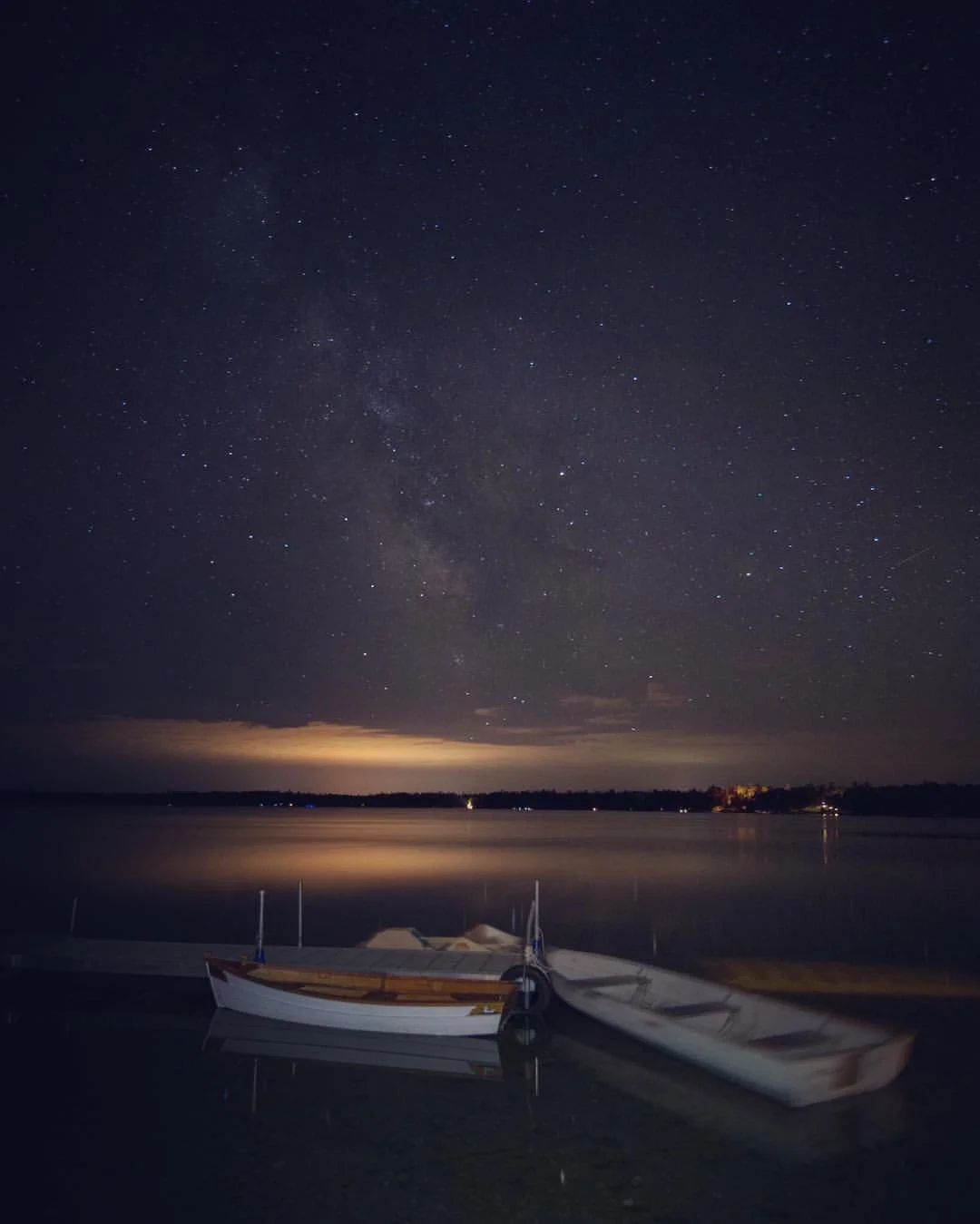 Nighttime scene over a lake with a star-filled sky and the Milky Way, two small boats floating on the calm water.