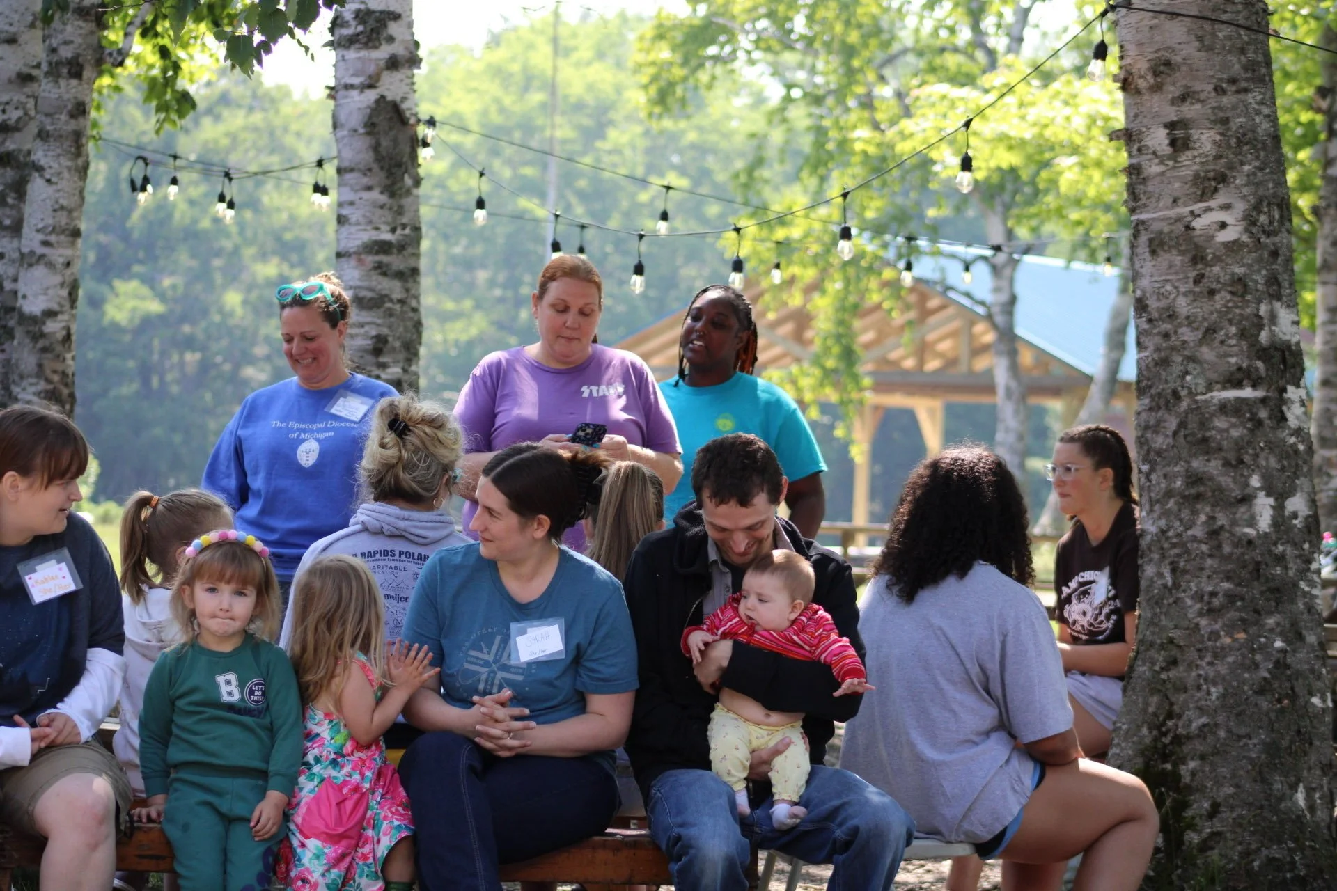 A group of people, including children and adults, sitting outdoors amongst trees, under string lights, at what appears to be a gathering or event during daytime.