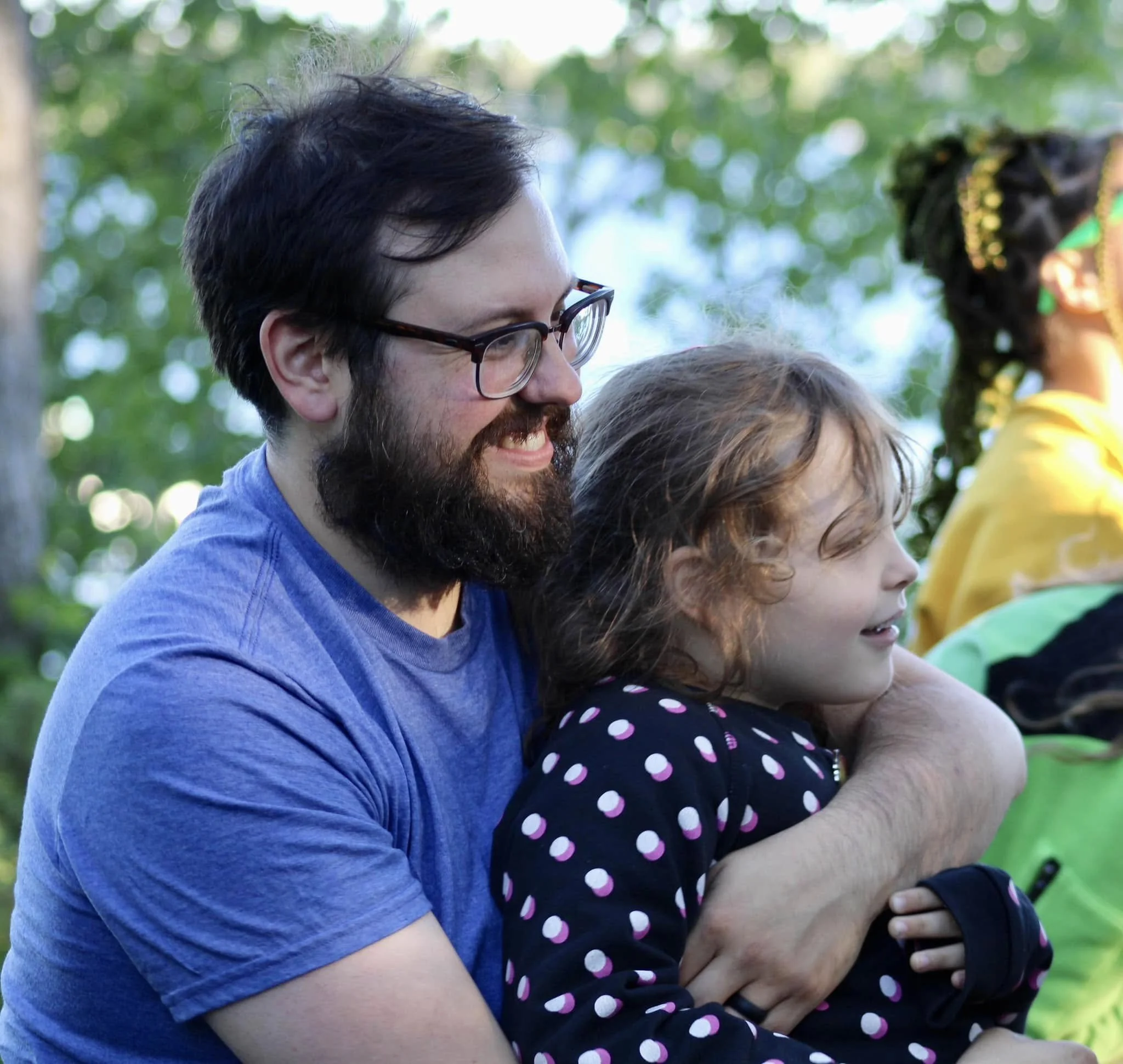 A man with glasses and a beard smiling while holding a young girl with curly hair in a polka dot jacket on his lap outdoors. There are trees and people in the background.