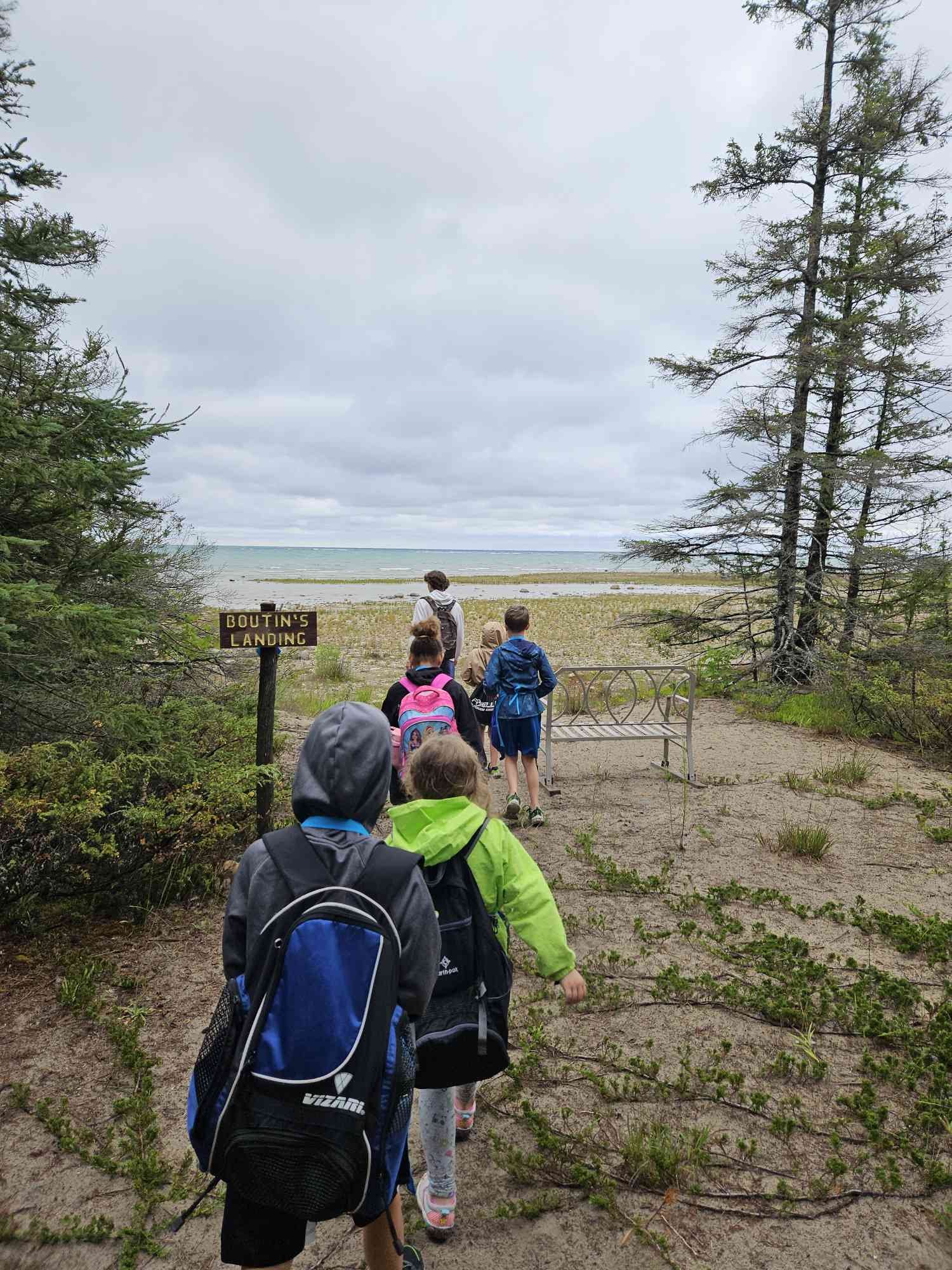 A group of children with backpacks walking towards a beach, with trees on both sides and cloudy sky, near a sign that says 'Boutins Landing'.