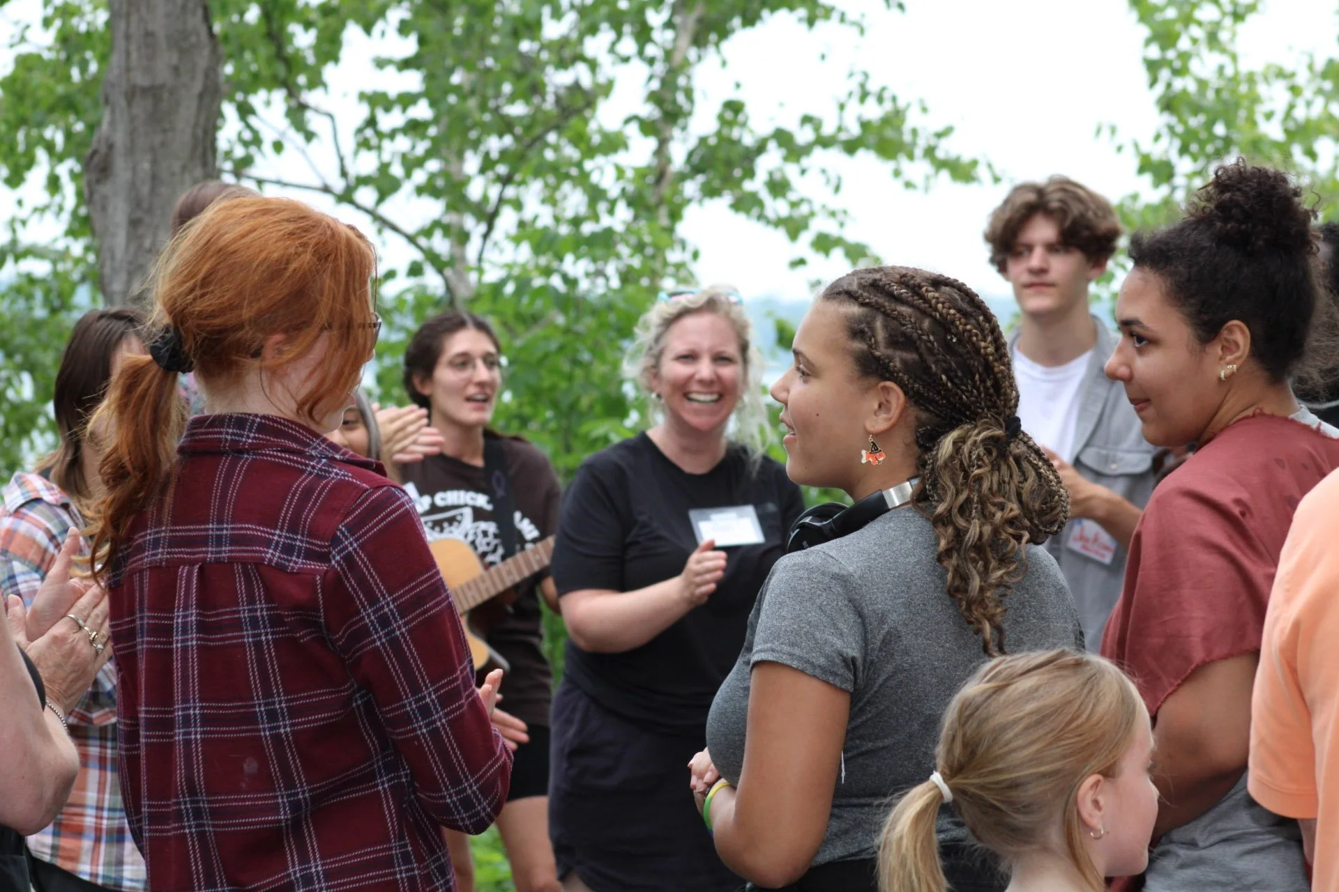 A group of people outdoors in a circle, smiling and engaging in conversation, with trees and green foliage in the background.