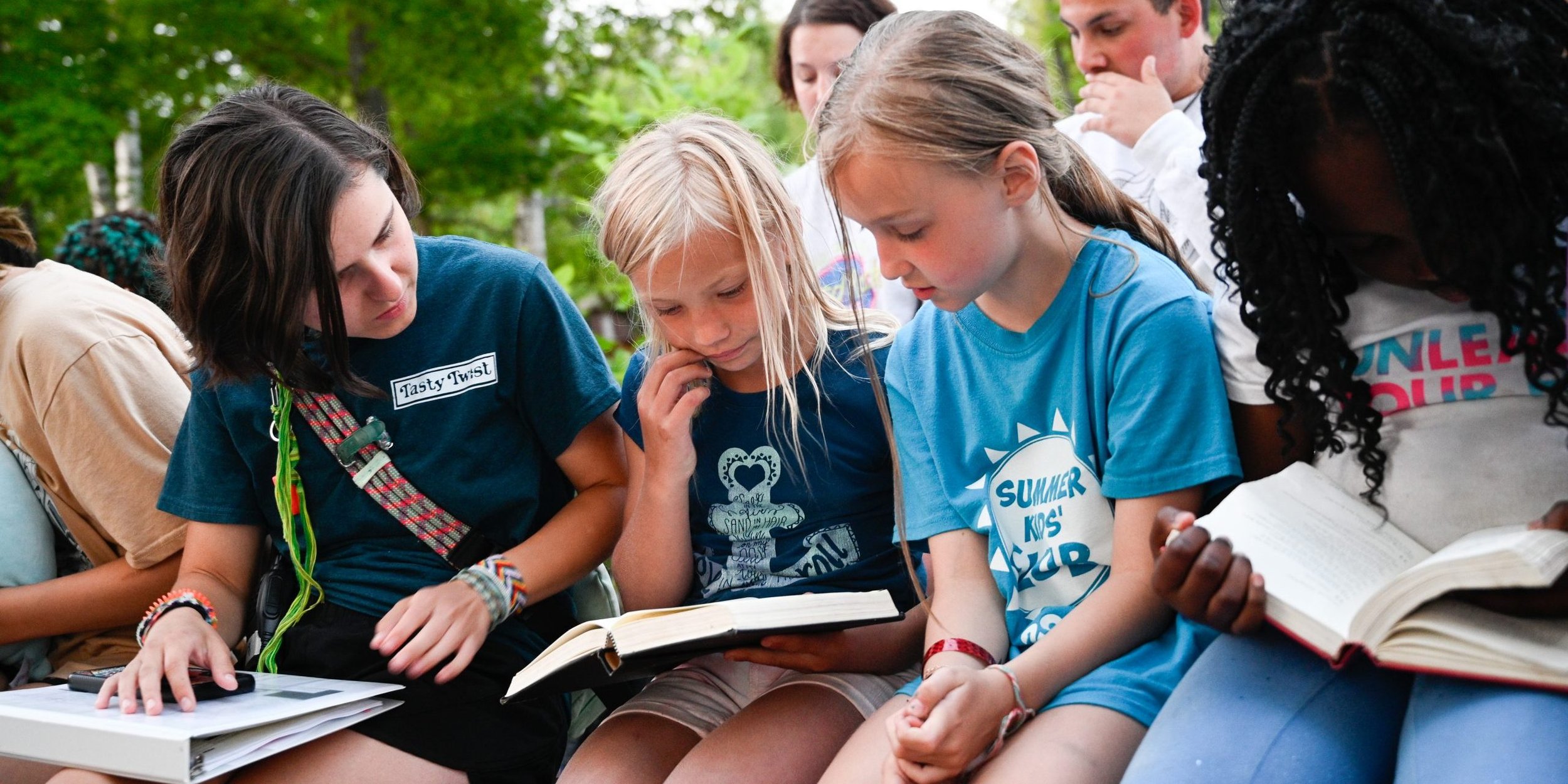 A group of children gathered outdoors, looking at a book together, surrounded by trees and greenery.