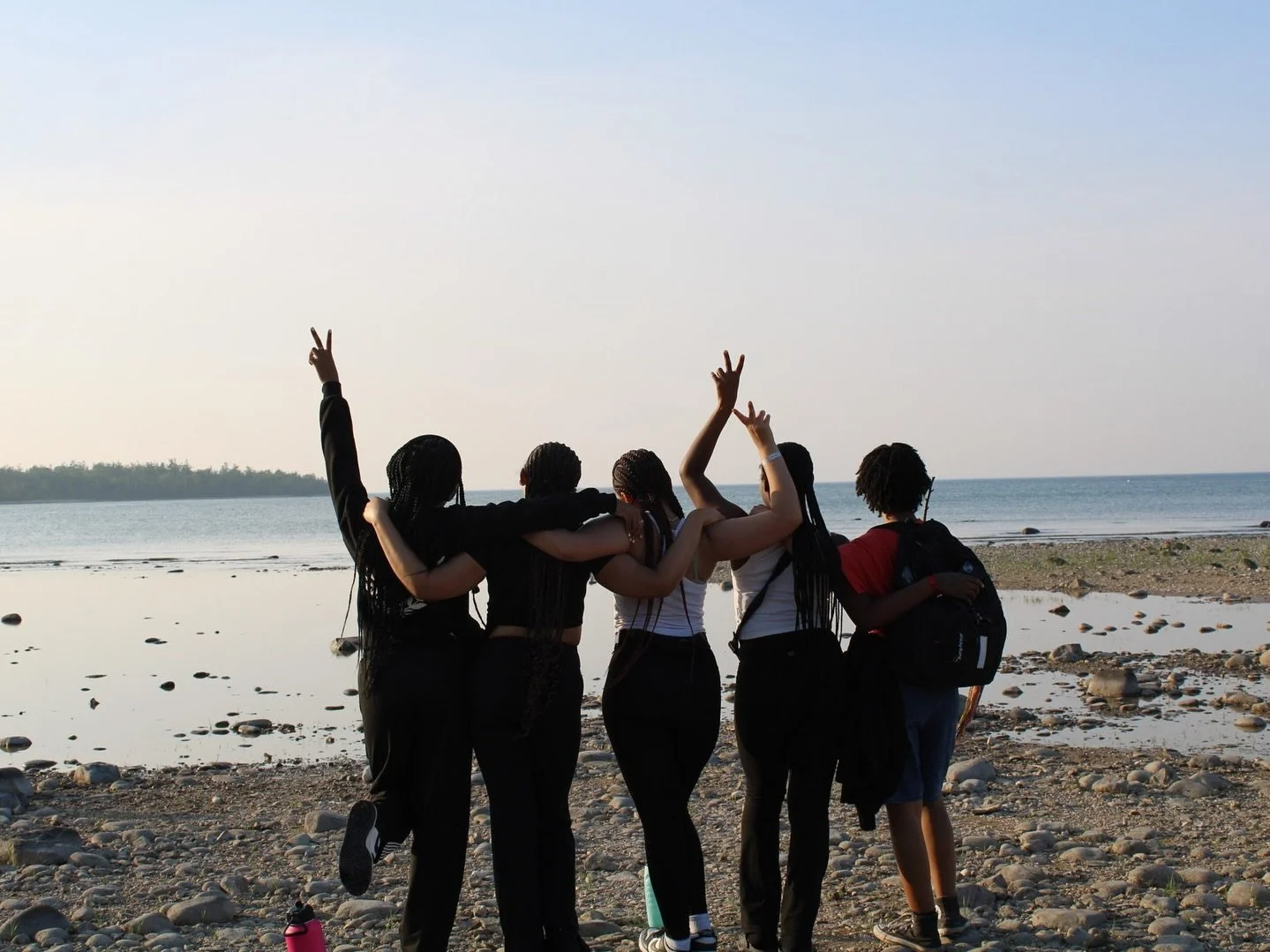 A group of five friends stands on a rocky beach with their arms around each other, facing the water, during sunset. Some are making peace signs, and one person has a backpack.