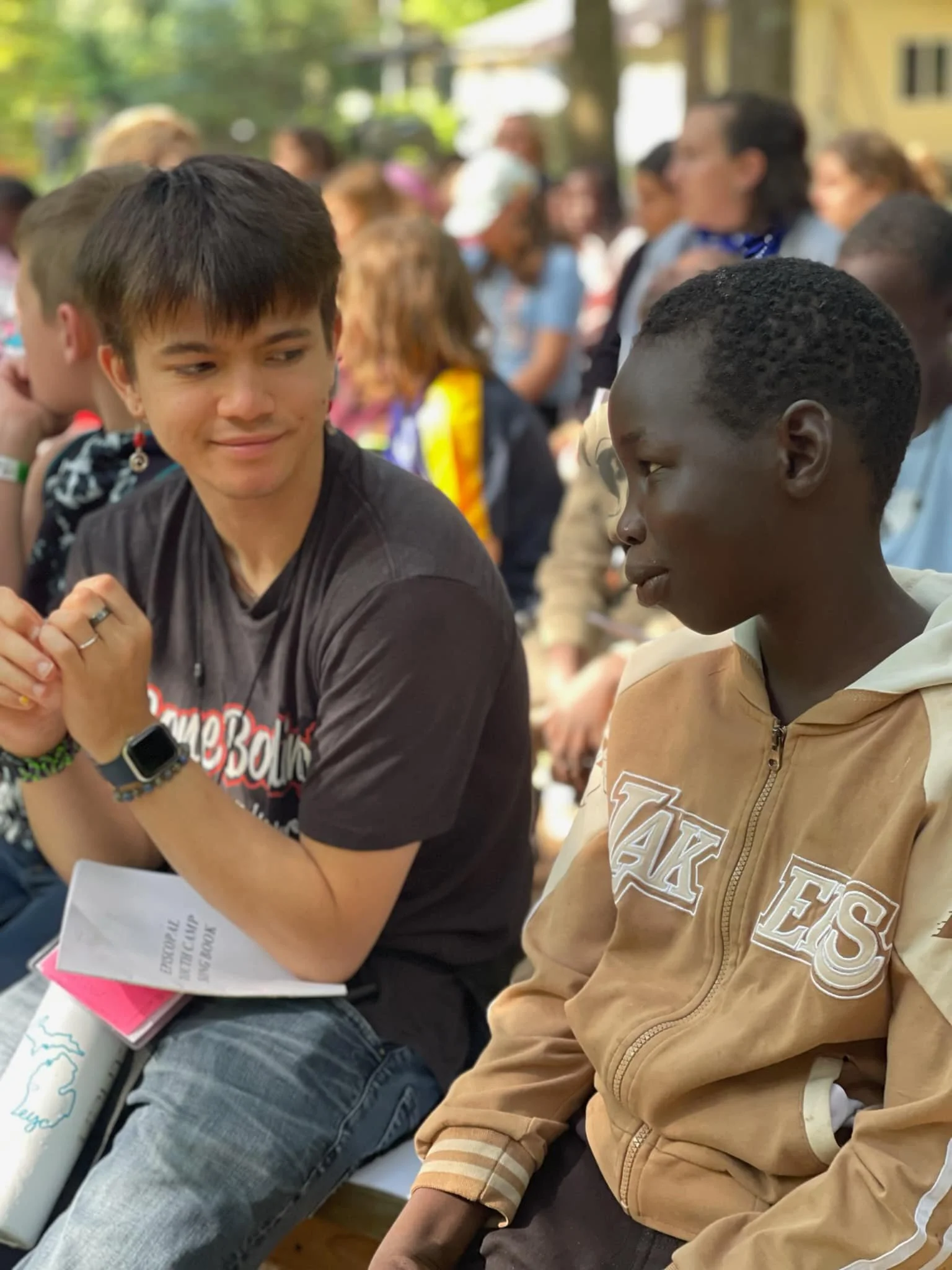 Two young males sitting outdoors, engaged in a conversation among a crowd of people at an event, with trees and colorful tents in the background.
