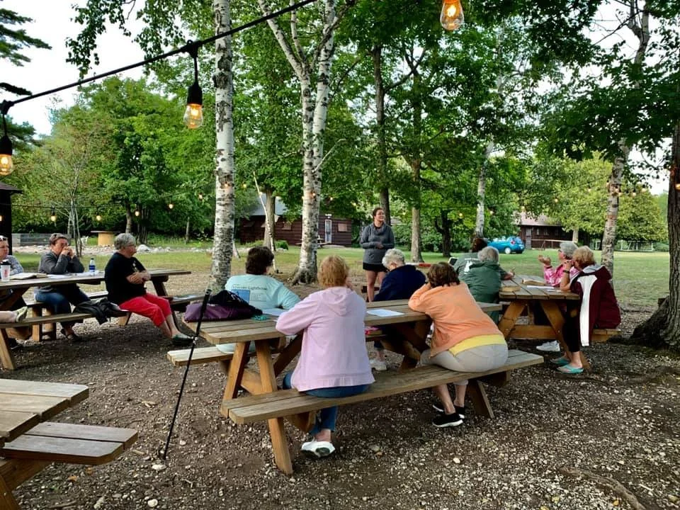 A group of people gathered outdoors around picnic tables in a wooded area, listening to a speaker standing in front of them. String lights hang overhead, and there are trees and a small building in the background.