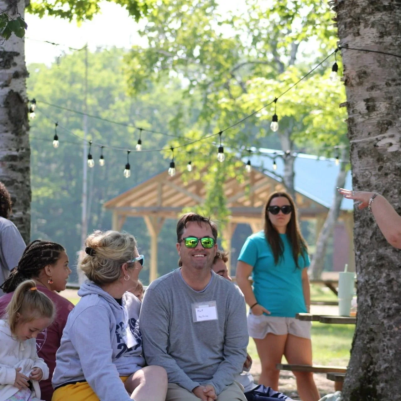 A group of people sitting outdoors under string lights and surrounded by trees, with a man wearing sunglasses smiling and a woman standing in the background.
