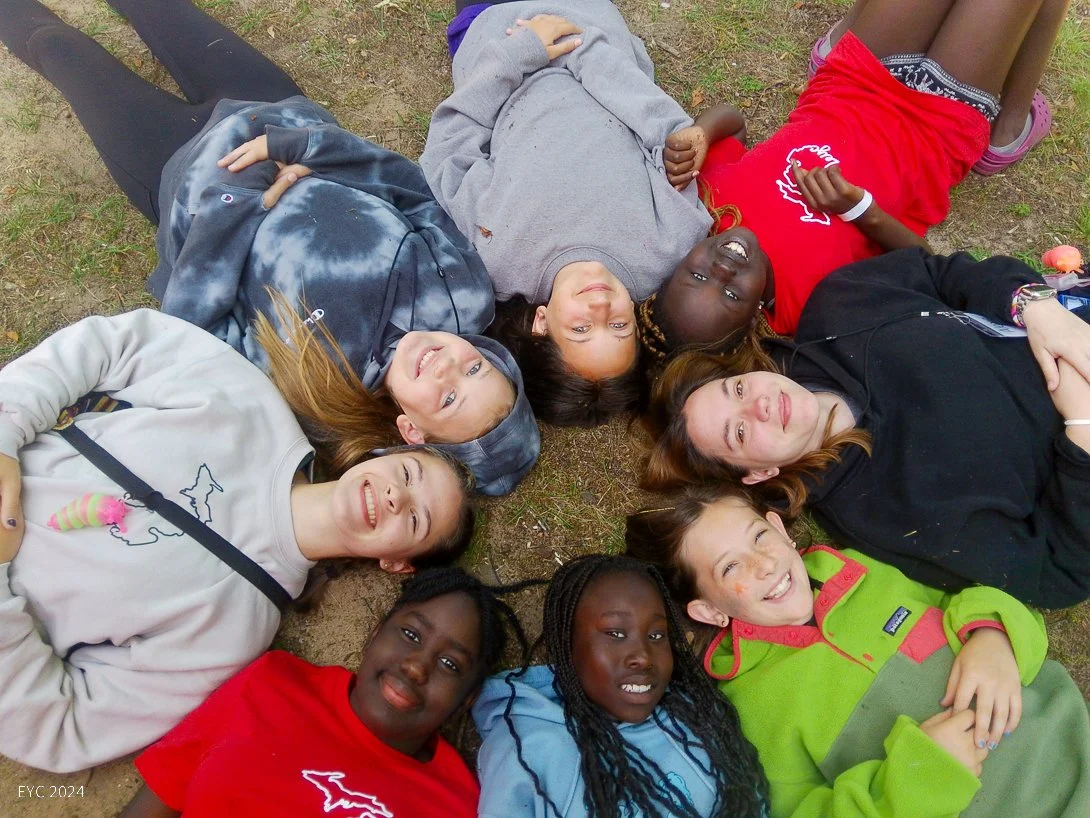 A group of children lying on the ground in a circle, looking up and smiling, outdoors on grass.
