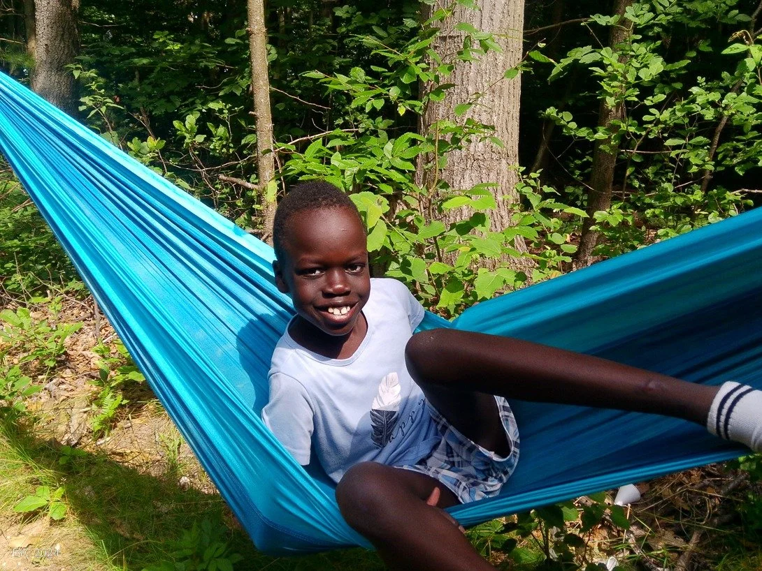 A young boy smiling and lying in a blue hammock outdoors surrounded by green trees and plants.