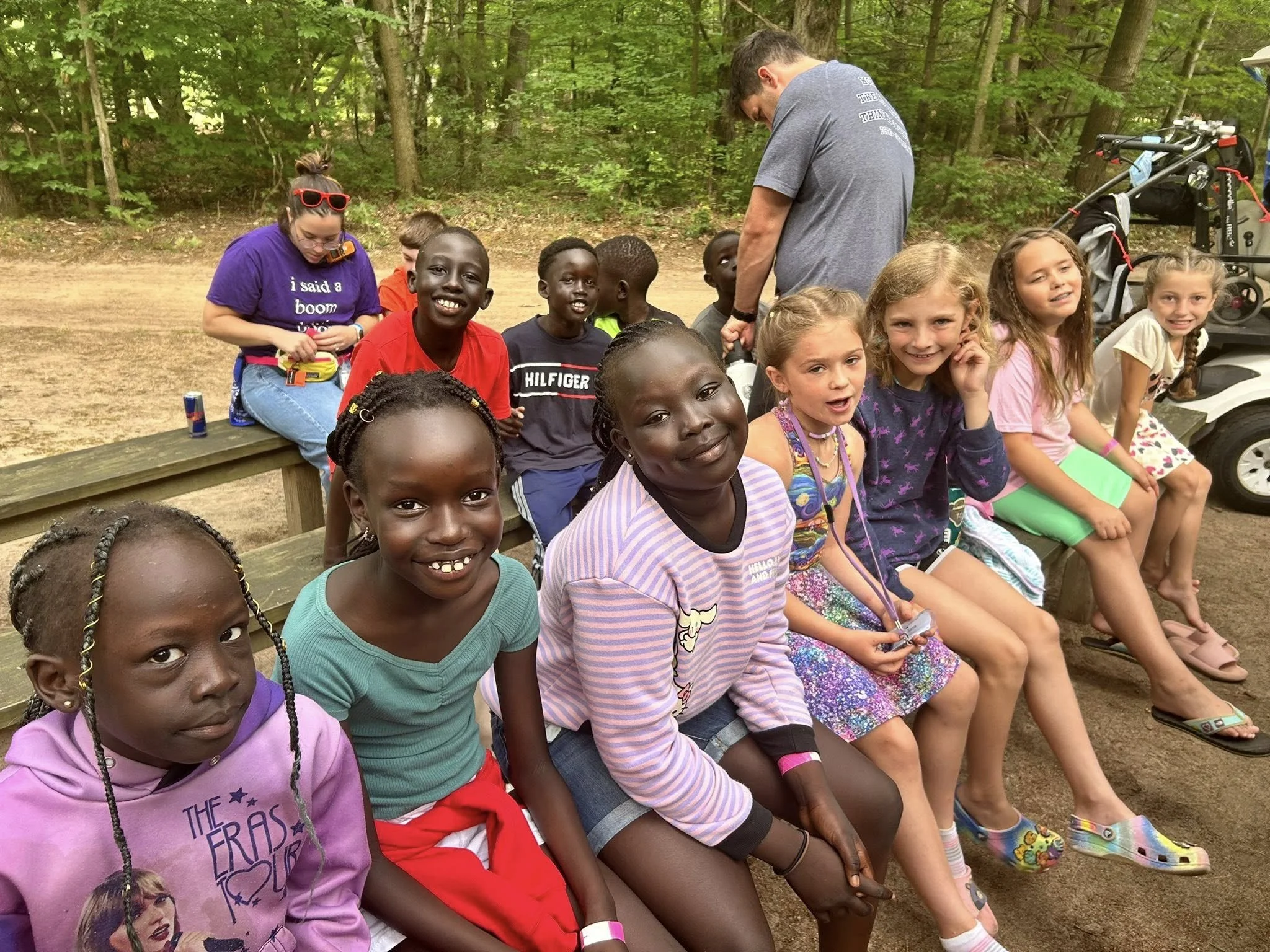 Group of children sitting on a wooden bench outdoors, with some smiling and others talking, surrounded by trees, with an adult standing behind them and a golf cart nearby.