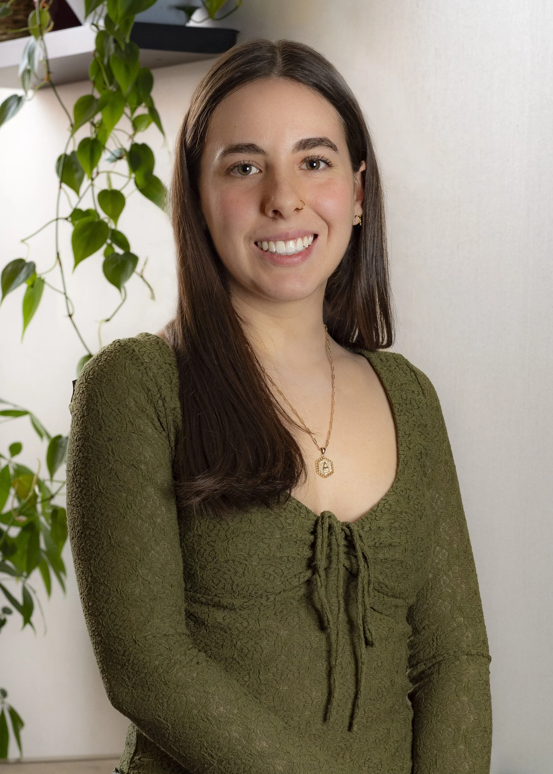A young woman with long brown hair, wearing an olive green textured top with a tie at the neckline, a gold necklace with a pendant, and small gold earrings, standing in front of a white wall and a plant with green leaves.