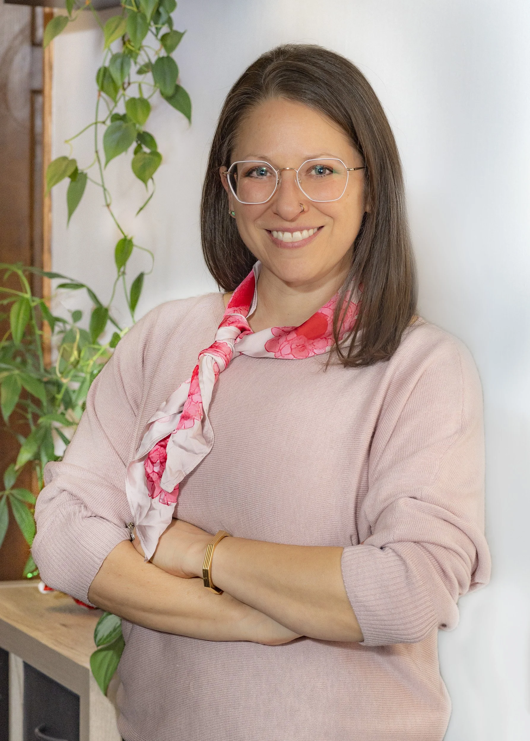 A woman with brown hair, glasses, and a pink sweater standing indoors near green houseplants, smiling with arms crossed.
