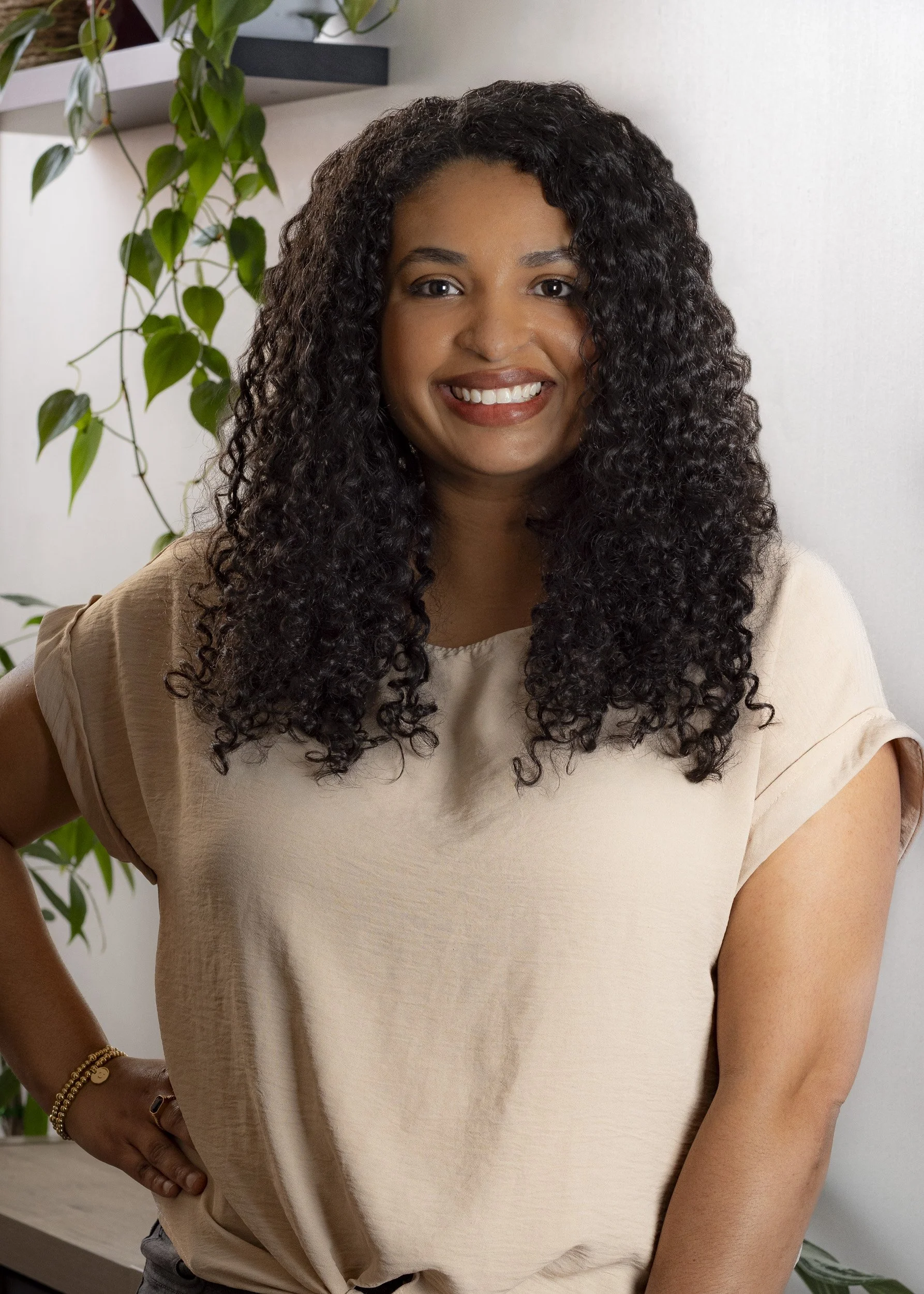 Smiling woman with dark curly hair wearing a beige top, standing indoors near a white wall and a green plant.