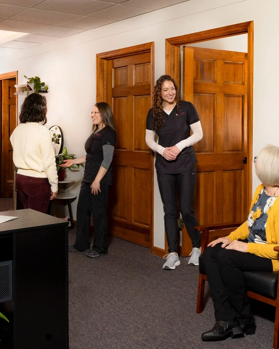 Four women in an office or waiting room, three standing and one sitting, engaging in conversation and smiling.