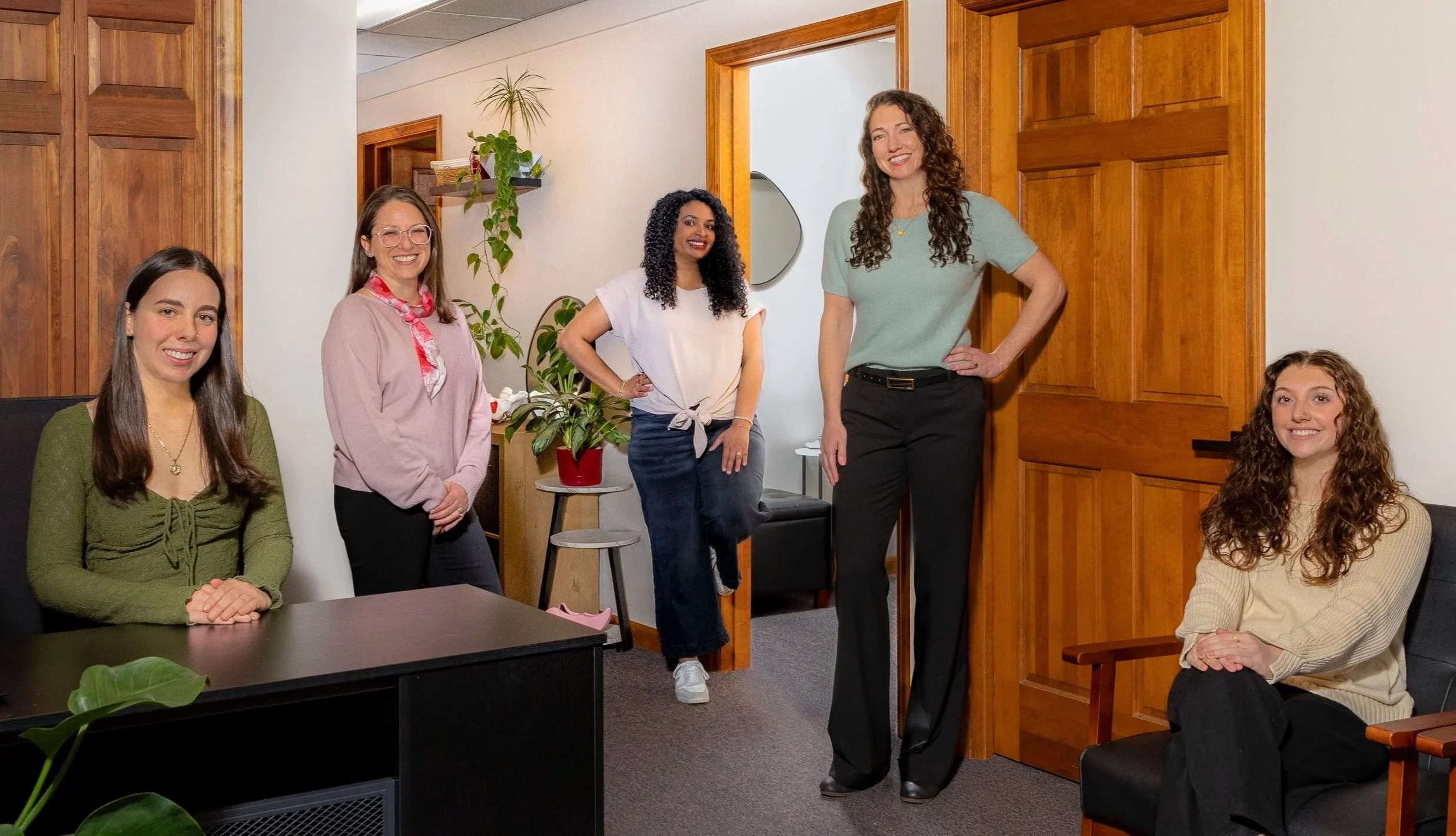 Five women in a casual office space. Two are seated, three are standing, smiling. The room has wood paneling, plants, and a mirror.