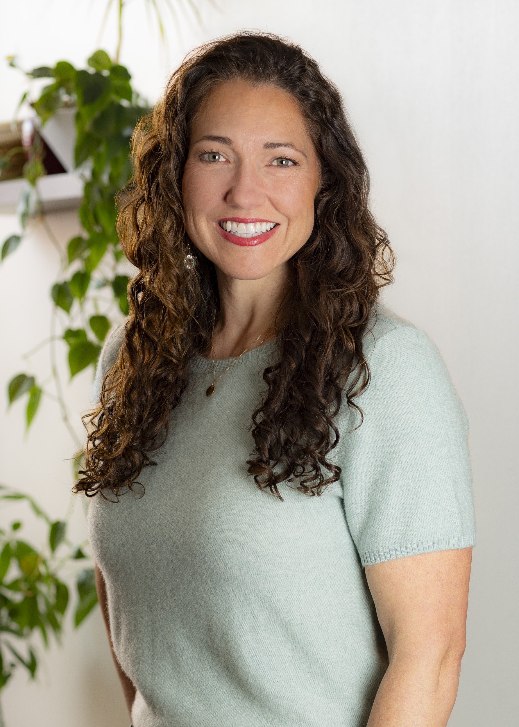 A woman with long, curly brown hair and fair skin, smiling, wearing a light green short-sleeved top, standing in front of a white wall and a green potted plant.