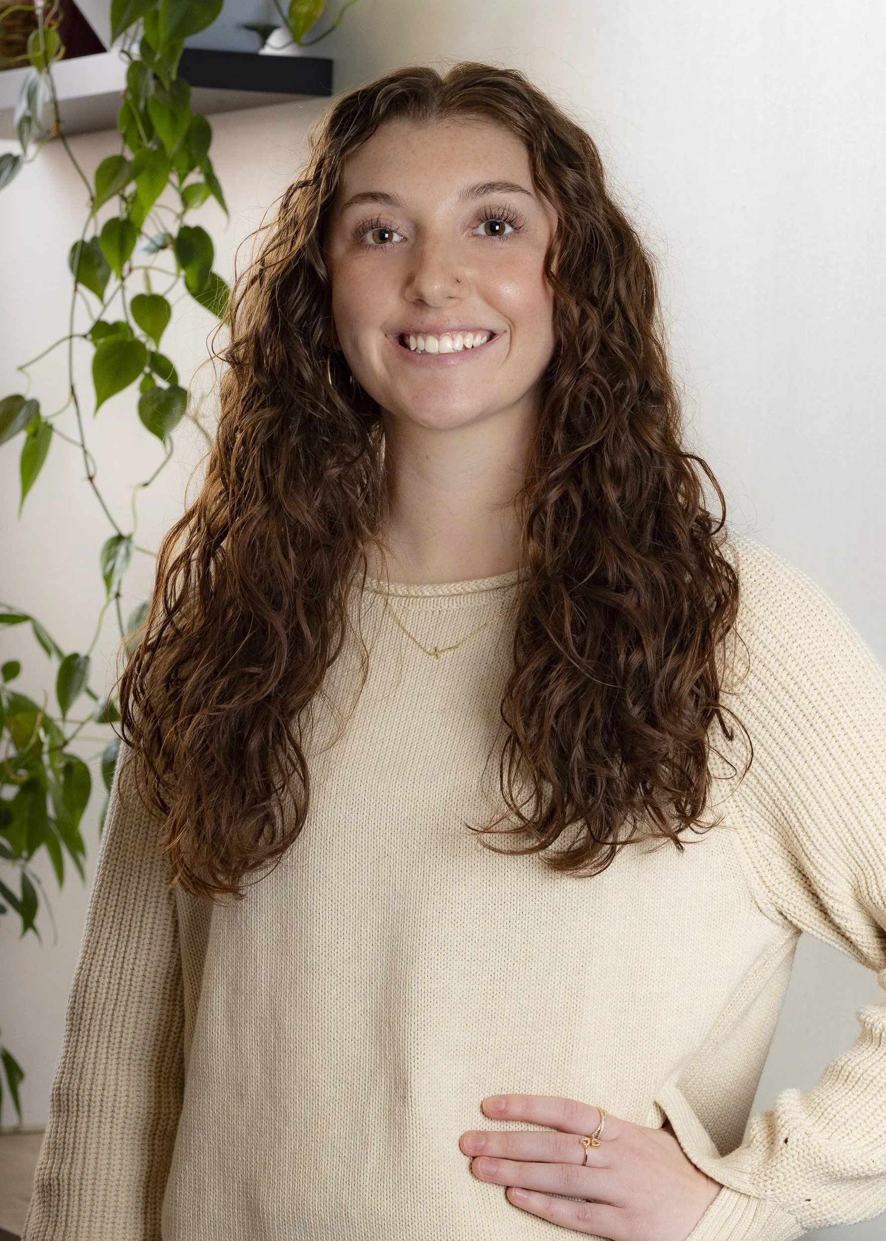 A young woman with long, curly brown hair, smiling and standing in front of a white wall with a green plant in the background.
