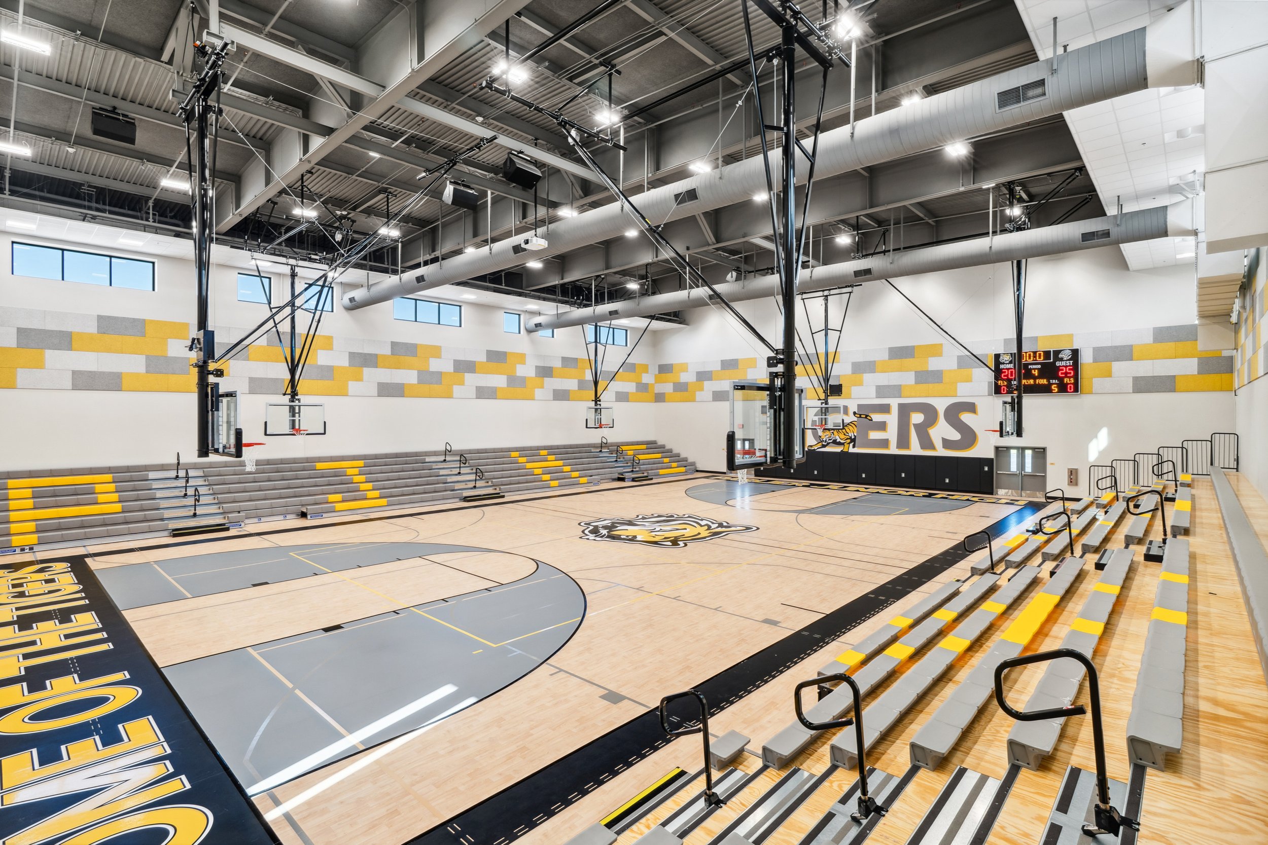 Inside a modern basketball gymnasium with a polished wooden court, multiple basketball hoops, and yellow and gray bleacher seating. The gym features high ceilings with large windows, industrial lighting, and a scoreboard at one end.