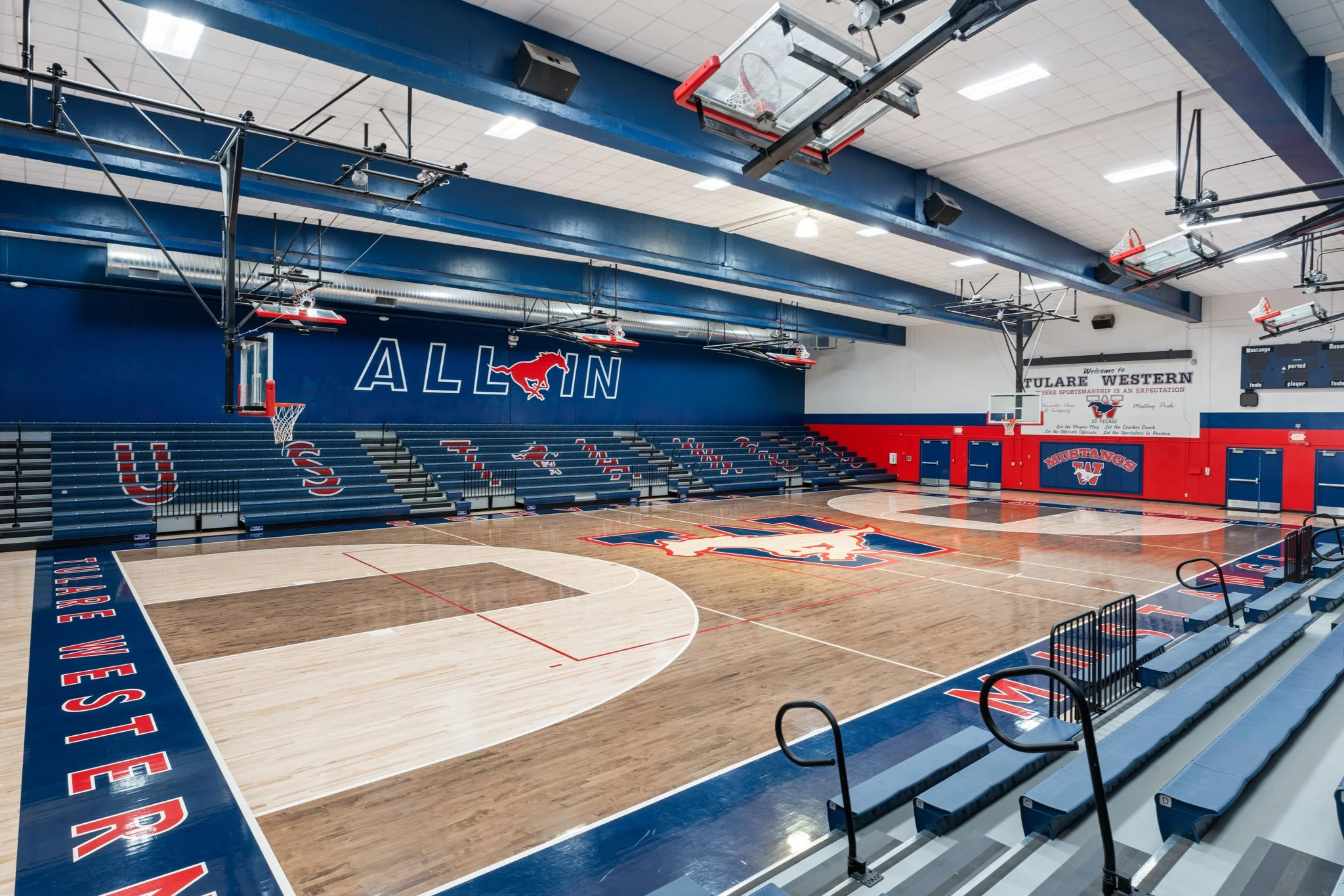 An indoor basketball court with a blue and red color scheme, featuring a large wall logo of a running mustang and the word 'ALLIN'; bleachers along one side; a basketball hoop with a net and backboard; and multiple ceiling-mounted lights and equipmen