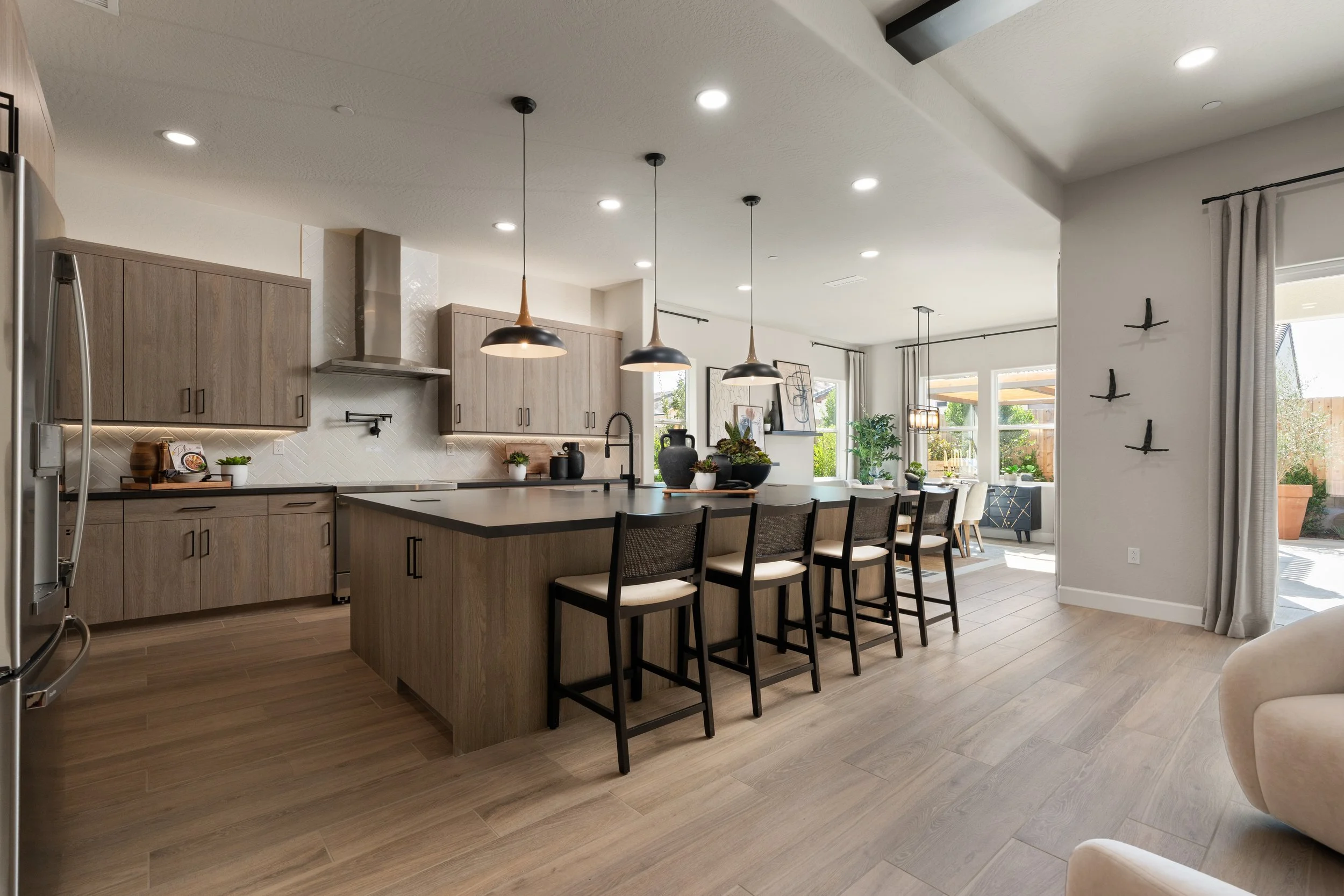 Open-concept kitchen and dining area with modern wooden cabinets, a large kitchen island with seating, hanging pendant lights, and large windows allowing natural light, decorated with plants and artwork.
