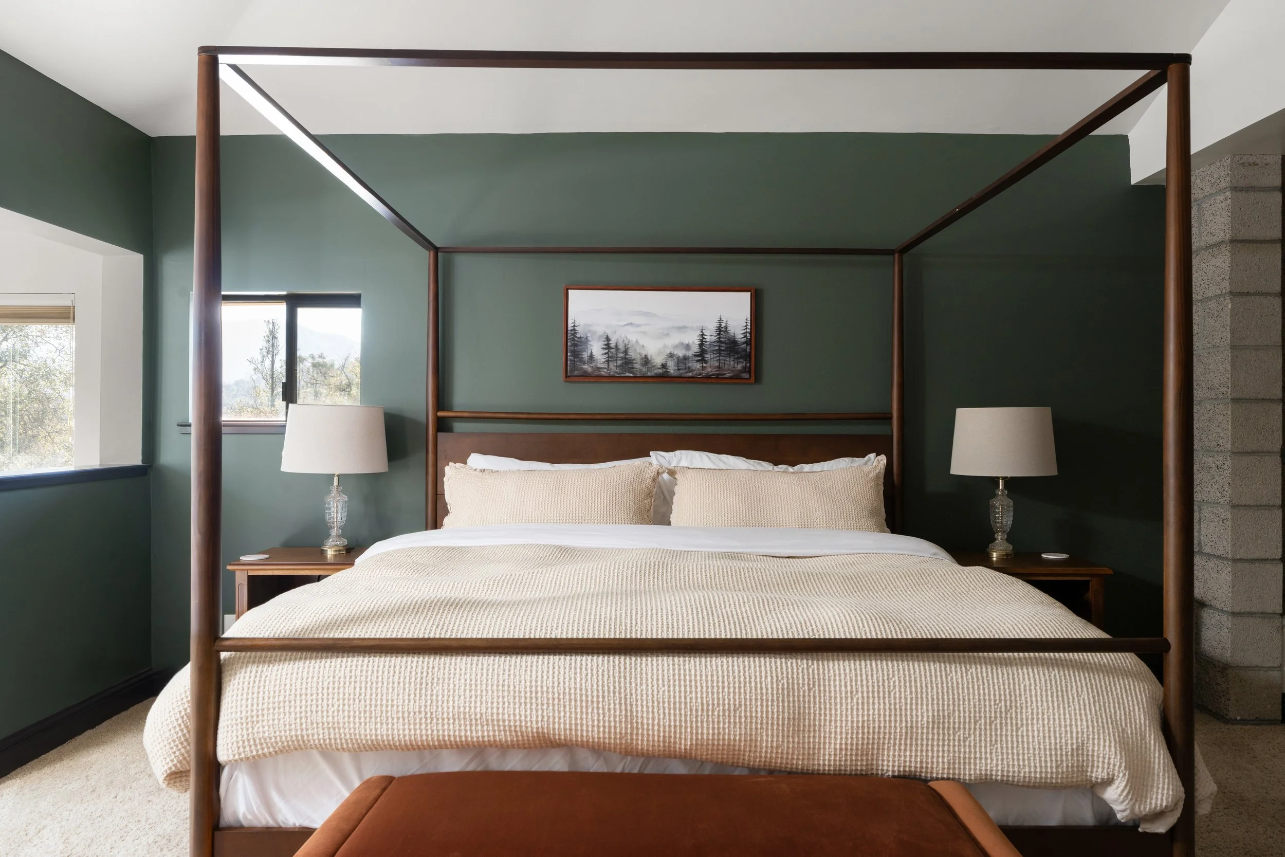A bedroom with a four-poster bed, beige bedding, and two white lamps on wooden nightstands. There is a framed landscape painting hanging above the bed, and windows with natural light coming in.