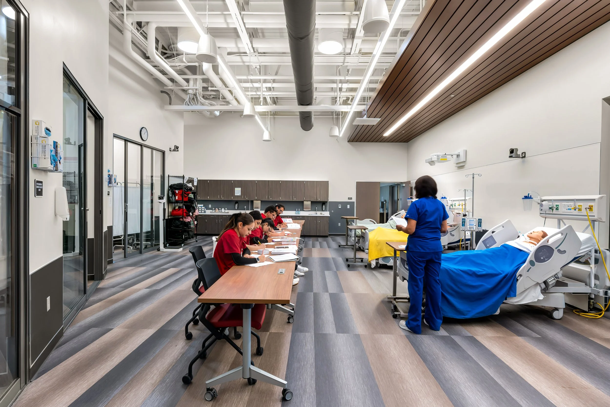 A hospital simulation room with medical mannequins in hospital beds, a nurse in blue scrubs standing by, and medical staff and students sitting at a table taking notes.