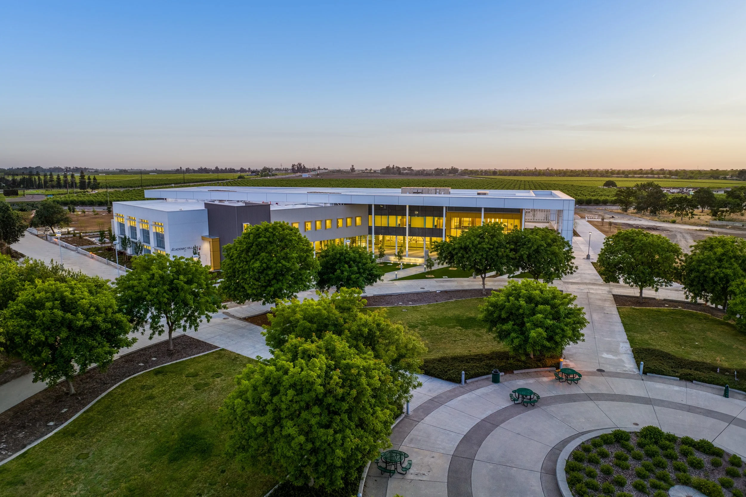 A modern educational building with large glass windows, situated in a green landscape with trees, a park, and open fields in the background during sunset.