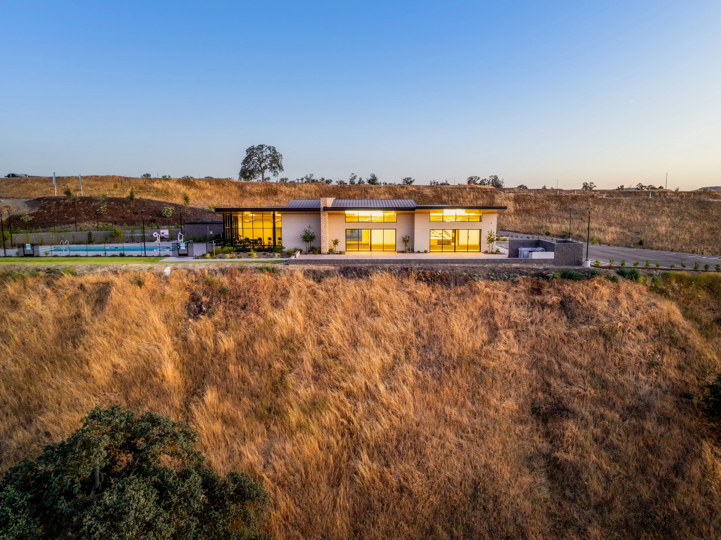 A modern house with large glass windows and a metal roof, surrounded by dry grass and sparse trees, during sunset.