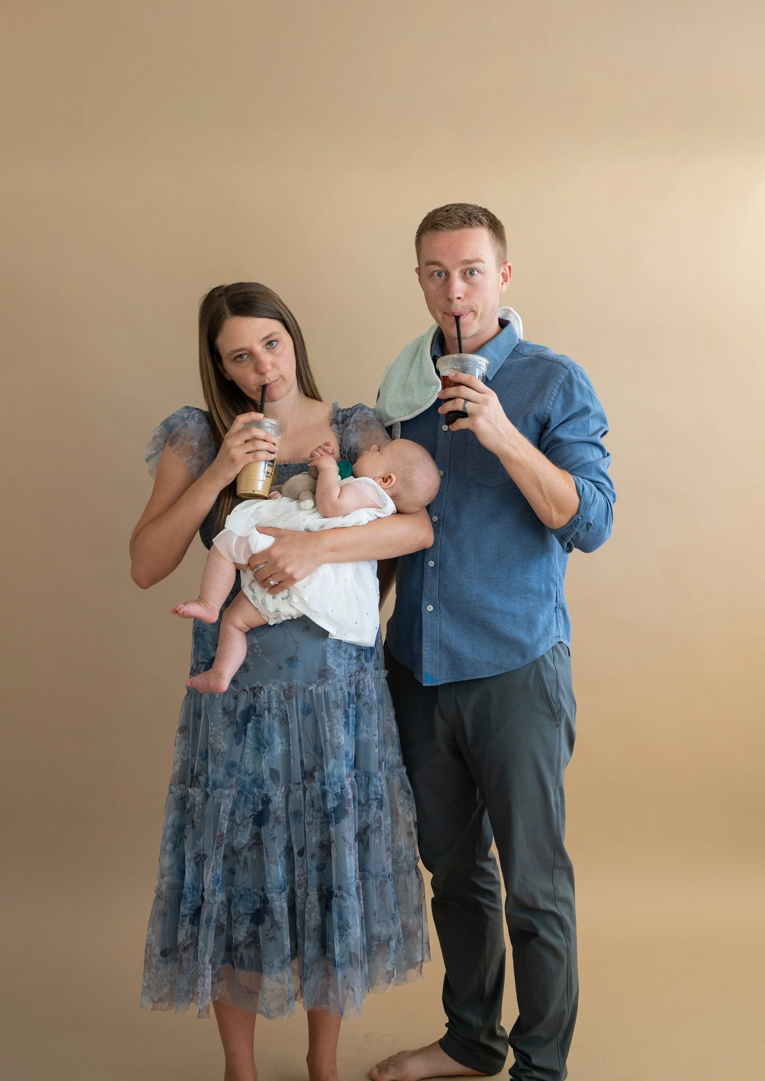 A woman holding a baby and a man drinking iced coffee with a bib around his neck, all standing against a beige wall.