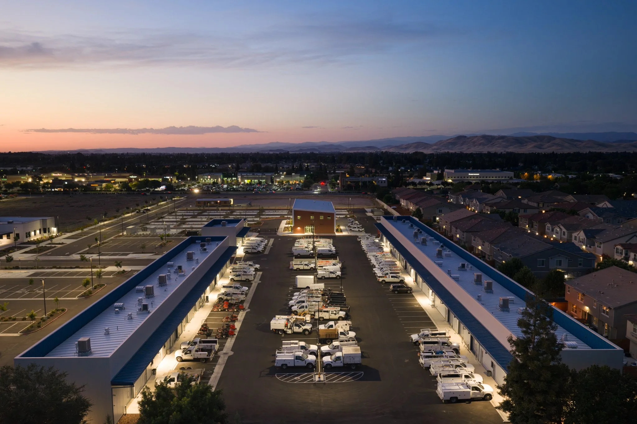 Nighttime view of a parking lot with many vehicles and a building, overlooking a suburban area with houses and a commercial area, hills in the background, and a partly cloudy sky with the remnants of sunset.