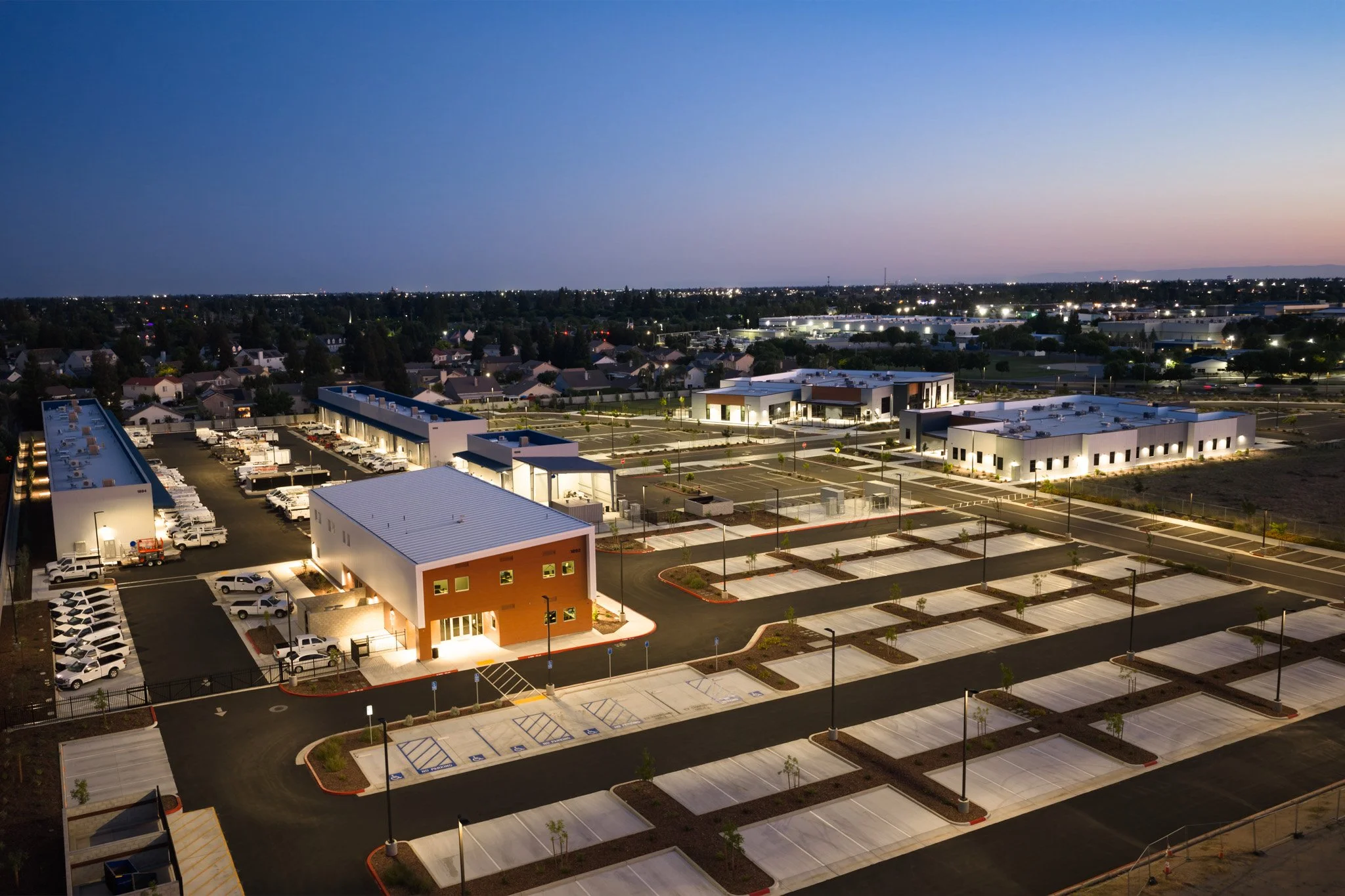 View of a commercial building parking lot at dusk with many empty parking spots, a few cars parked near the building, and a cityscape in the distance under a clear sky.
